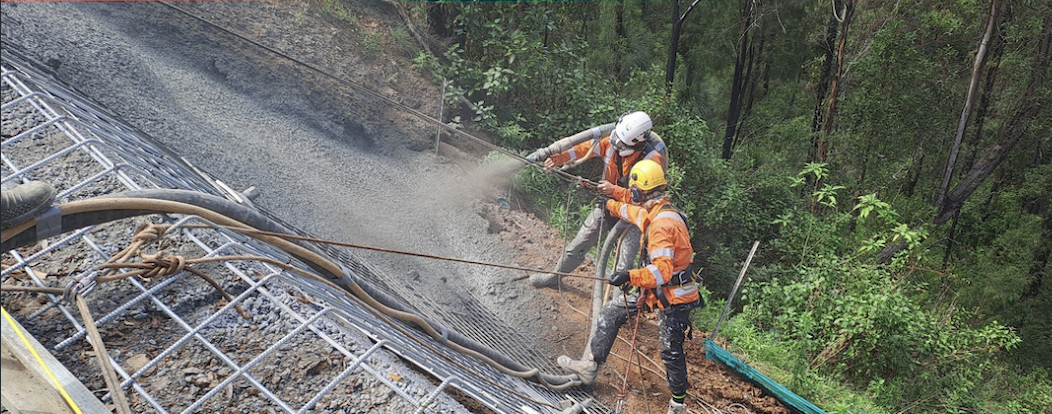 Two construction workers in safety gear working on a slope, securing a wire mesh for erosion control or slope stabilization in a forested area.