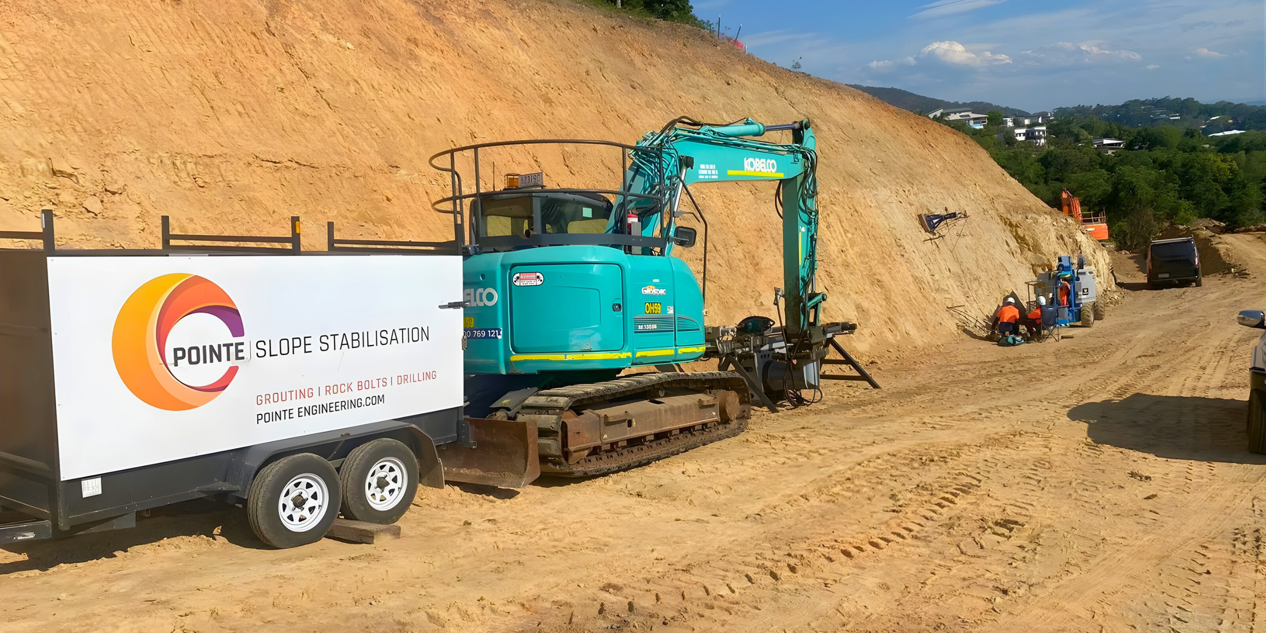 Construction site with a blue excavator and workers on a dirt slope, with cars parked nearby, under a partly cloudy sky.
