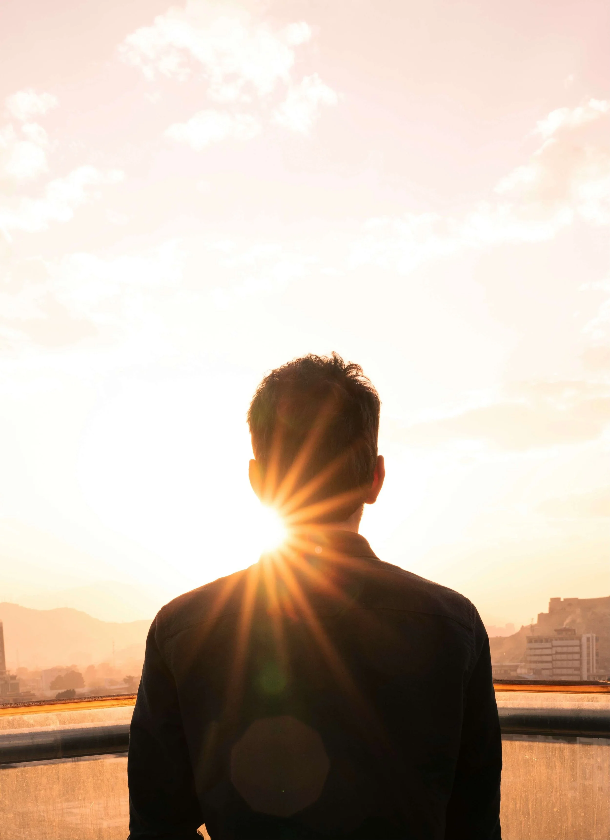 A person seen from behind standing outdoors during sunset, with sunlight shining behind their head, against a sky with clouds and a cityscape in the distance.