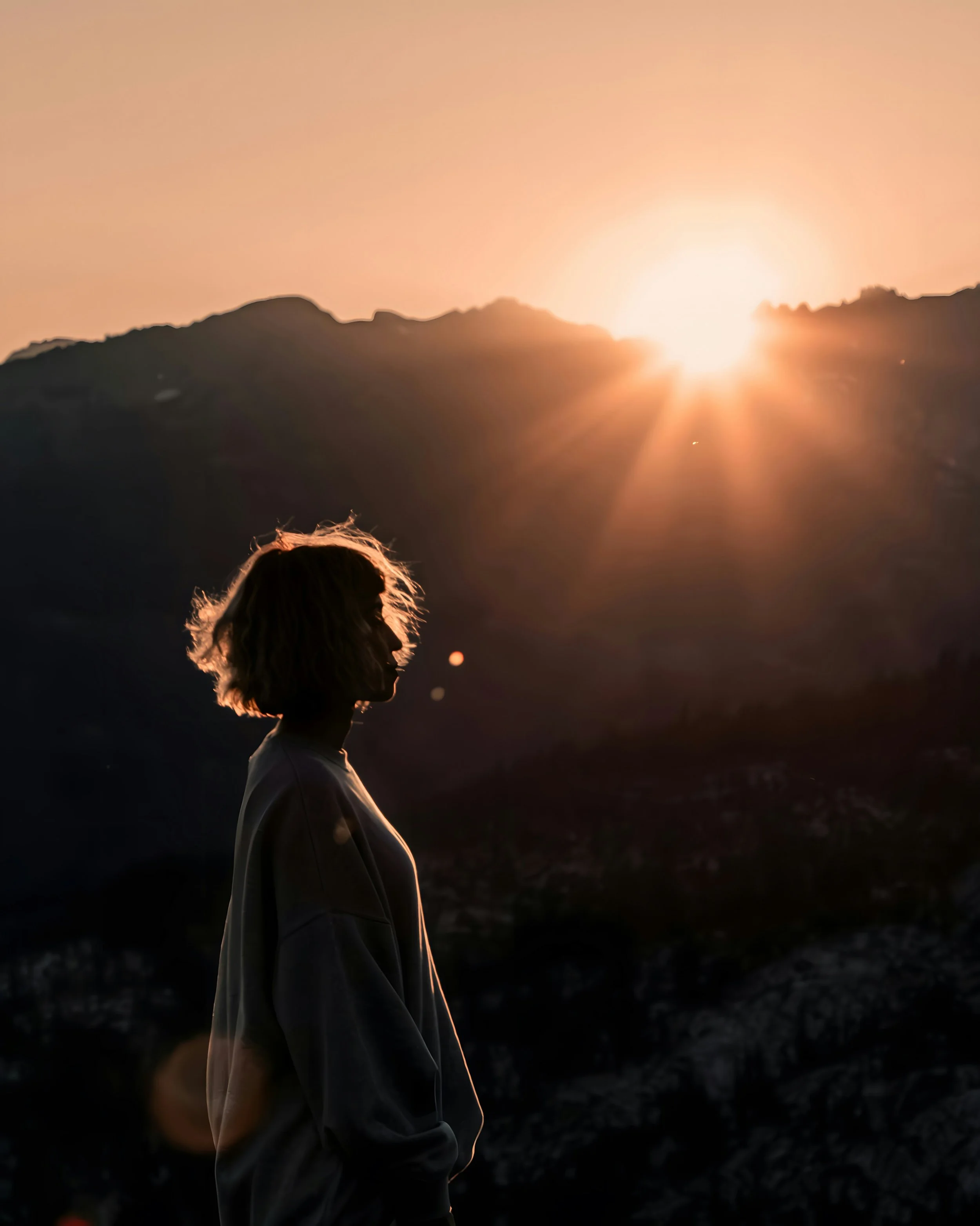 Silhouette of a woman with wavy hair wearing a light jacket, standing outdoors at sunset, with mountains visible in the background.