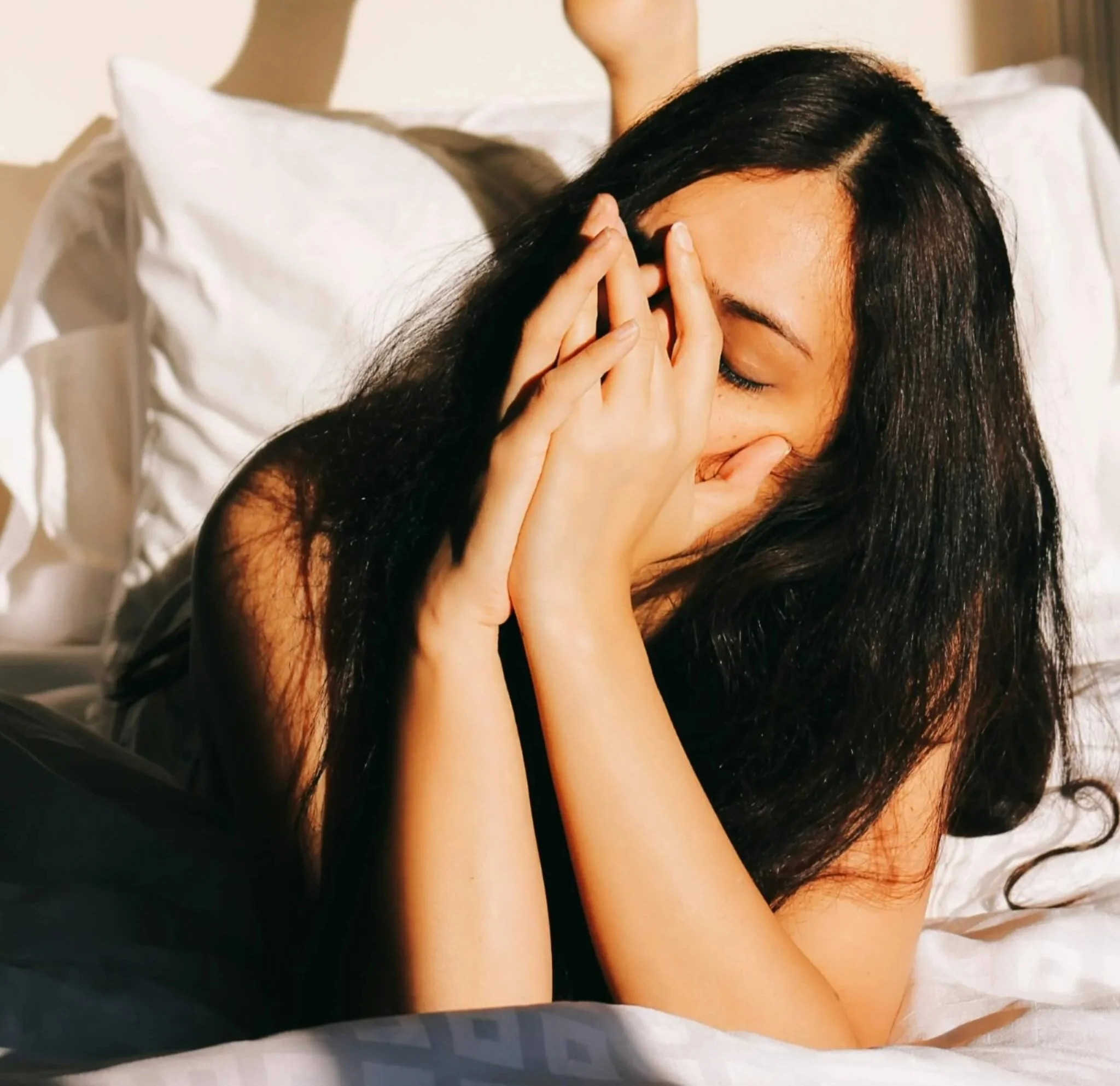A woman with long dark hair sitting on a bed, holding her head with her hands, appearing stressed or overwhelmed.
