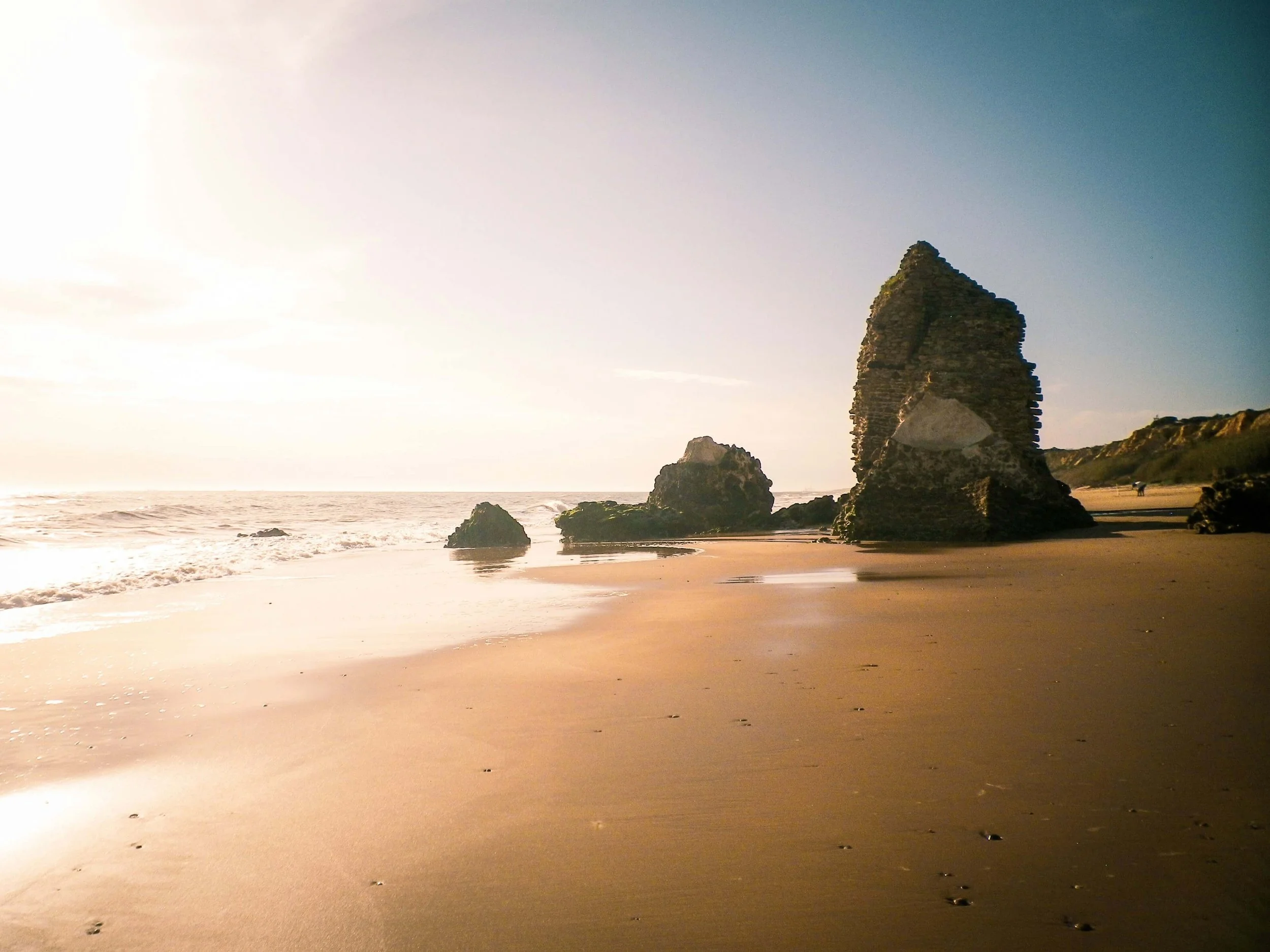 Beach with rocks and a tall sea stack during sunset, with the ocean in the background and a person walking along the shore.