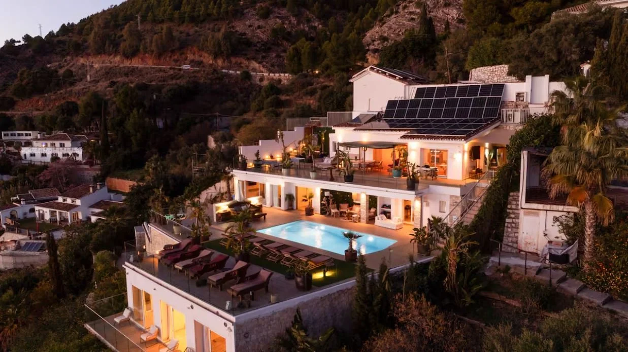 Luxury hillside house with outdoor pool, multiple decks, and solar panels at dusk surrounded by trees and neighboring homes