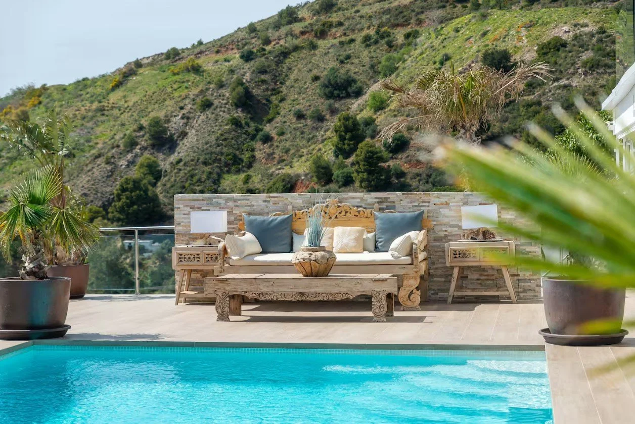 Outdoor patio with swimming pool, wooden couch with pillows, potted plants, and a hillside in the background.