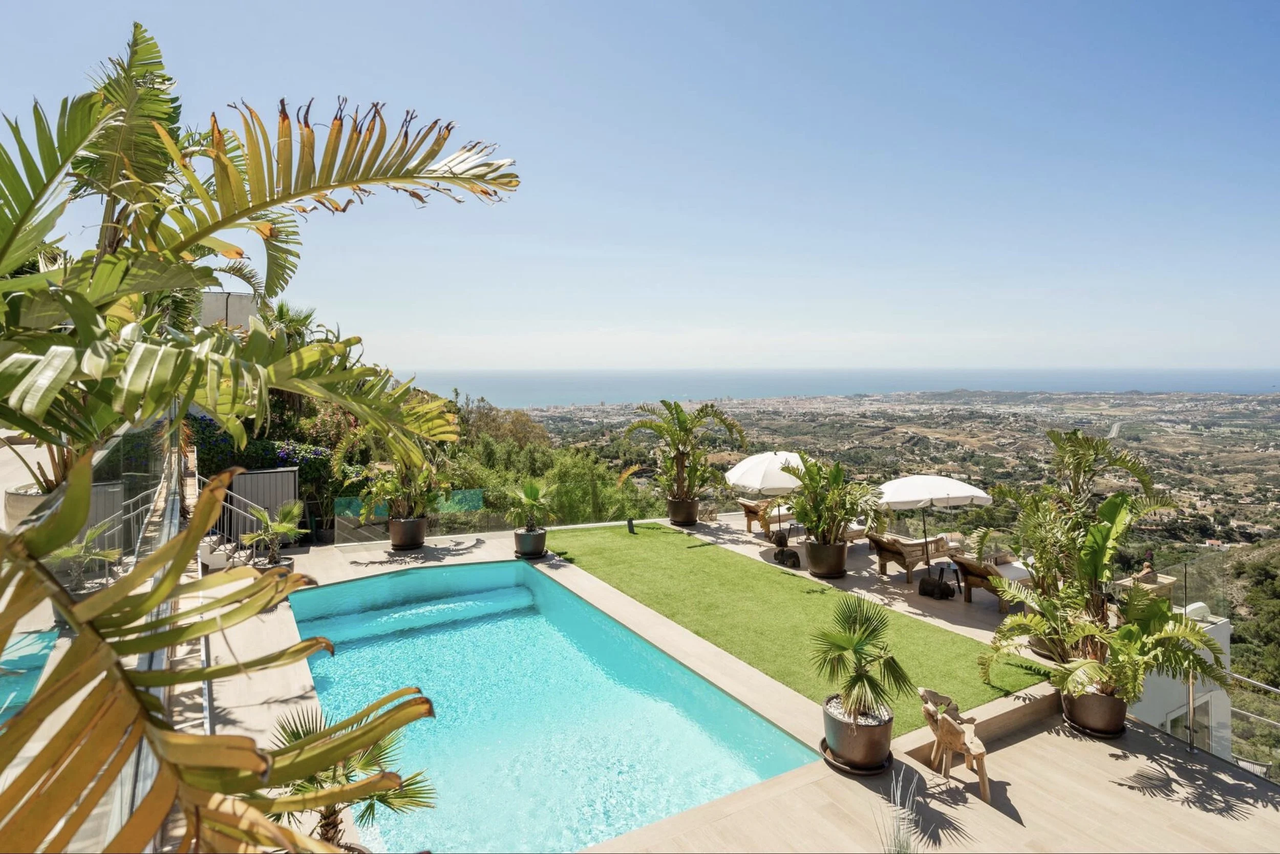 View of a swimming pool on a hillside with lush plants and trees, overlooking a city and coastline in the distance under a clear blue sky.