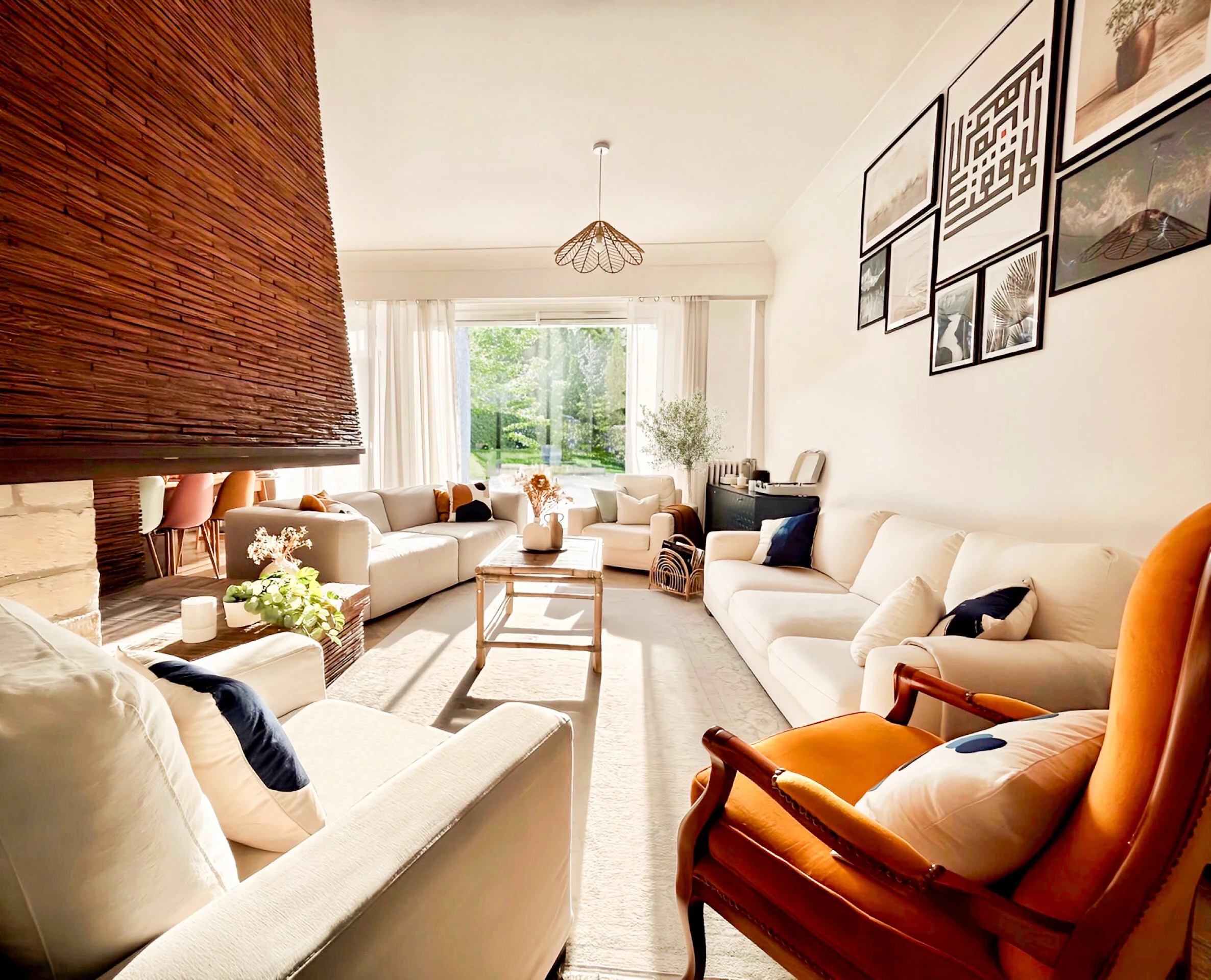 Bright living room with white sofas, an orange armchair, a wooden coffee table, large window with white curtains, greenery outside, wall art, and plants.