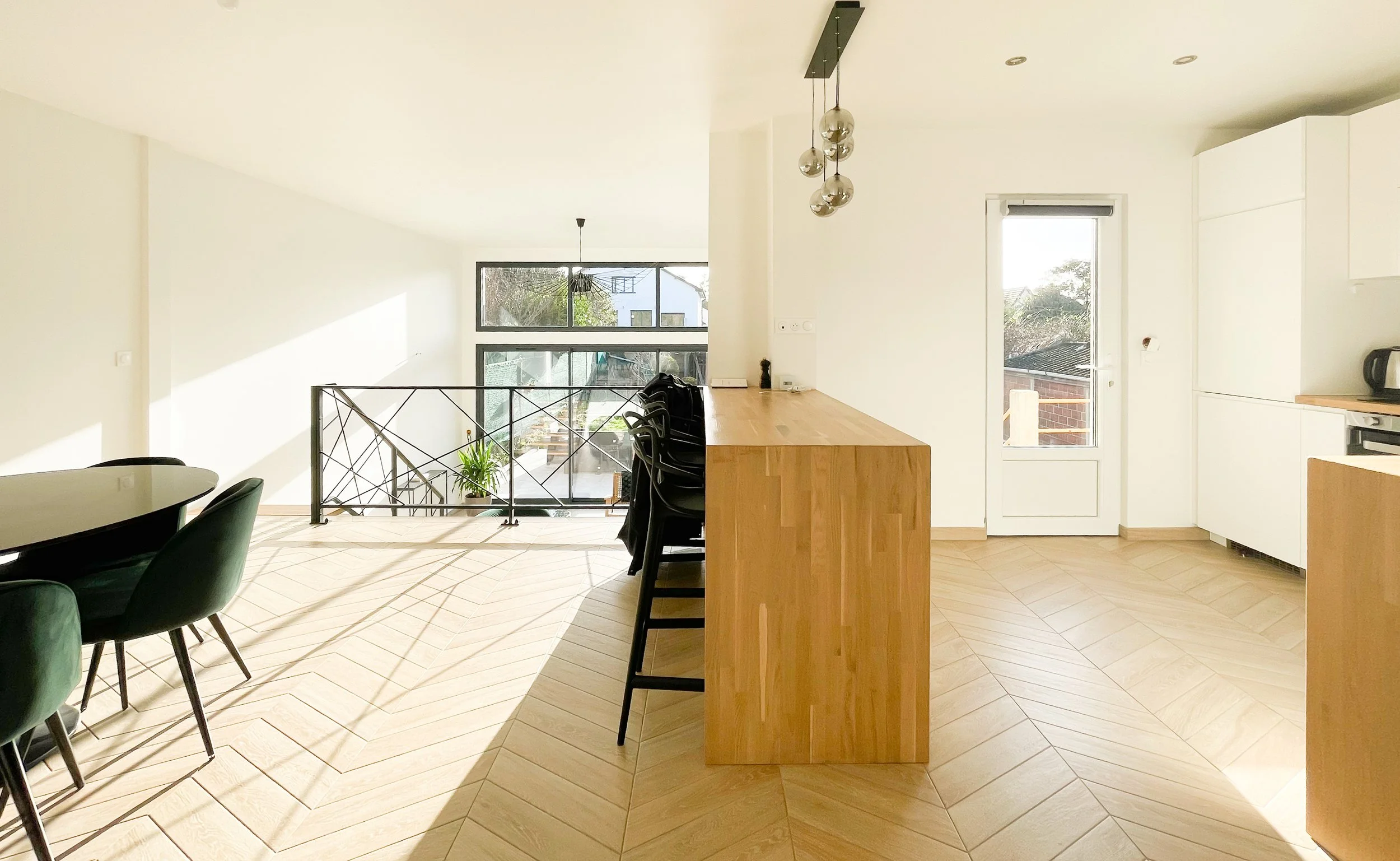 Bright, minimalist open-concept kitchen and dining area with wooden floor, white cabinets, a bar-height wooden countertop, black chairs, a glass door, and large windows bringing in natural light.