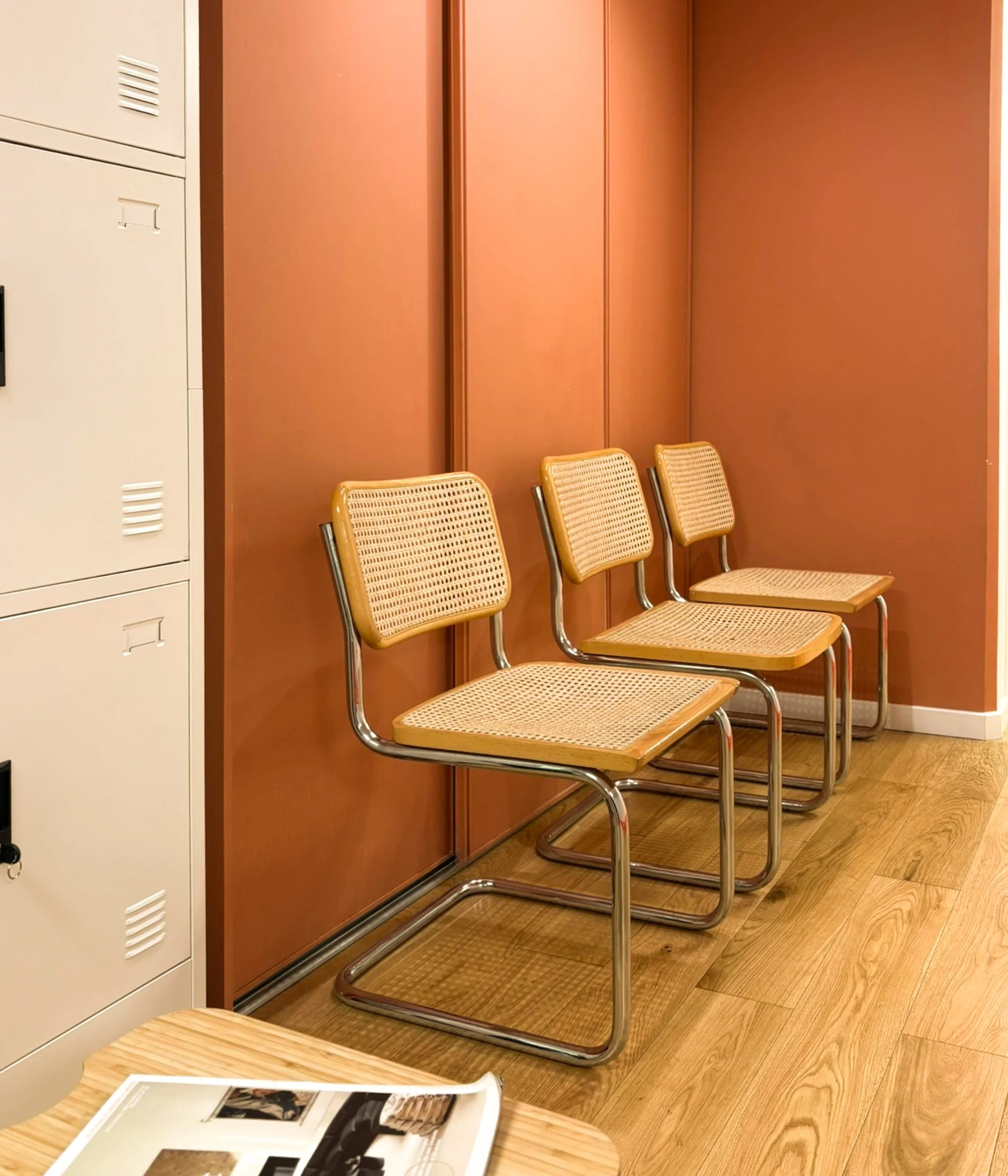 Three yellow cane and metal chairs aligned against a burnt orange wall in a waiting area with lockers and a wooden floor.