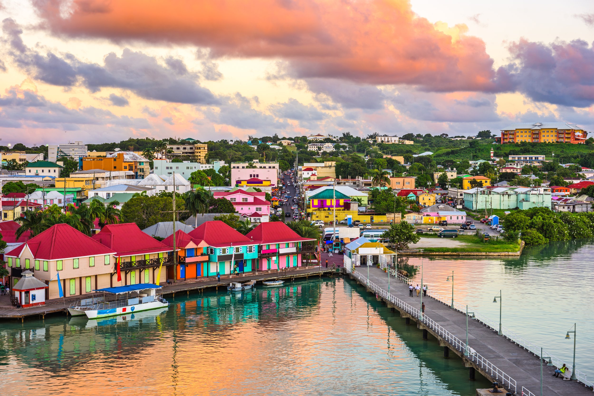 Colorful buildings along a waterfront with a pier extending into the water at sunset, with a sky of pink, orange, and purple clouds and rolling green hills in the background.