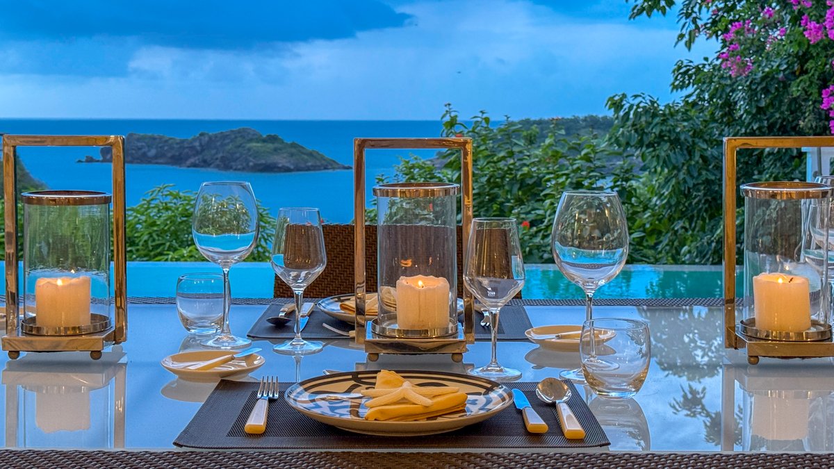 Elegant outdoor dining table set with candles, glasses, and cutlery, overlooking a scenic ocean with islands and lush greenery in the background.