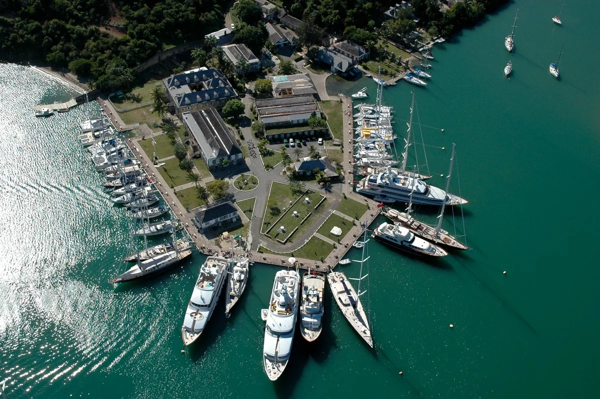 Aerial view of a marina with yachts docked along the piers and boats in the water, with buildings and trees surrounding the area.