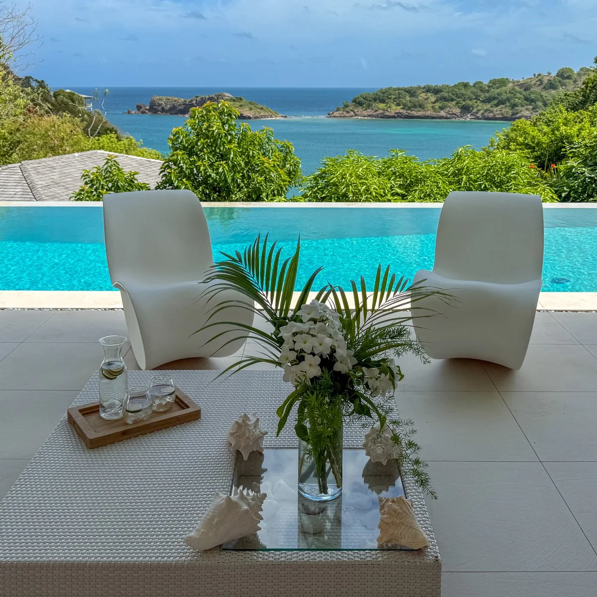 Luxury outdoor patio overlooking a swimming pool and ocean, with white chairs, a glass table with flowers, and lush green trees.