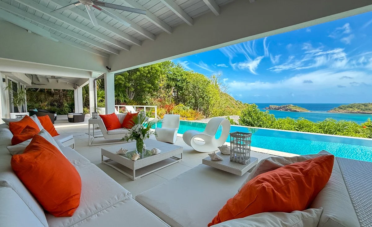 Luxury outdoor living space with white furniture and orange cushions overlooking ocean and lush greenery.
