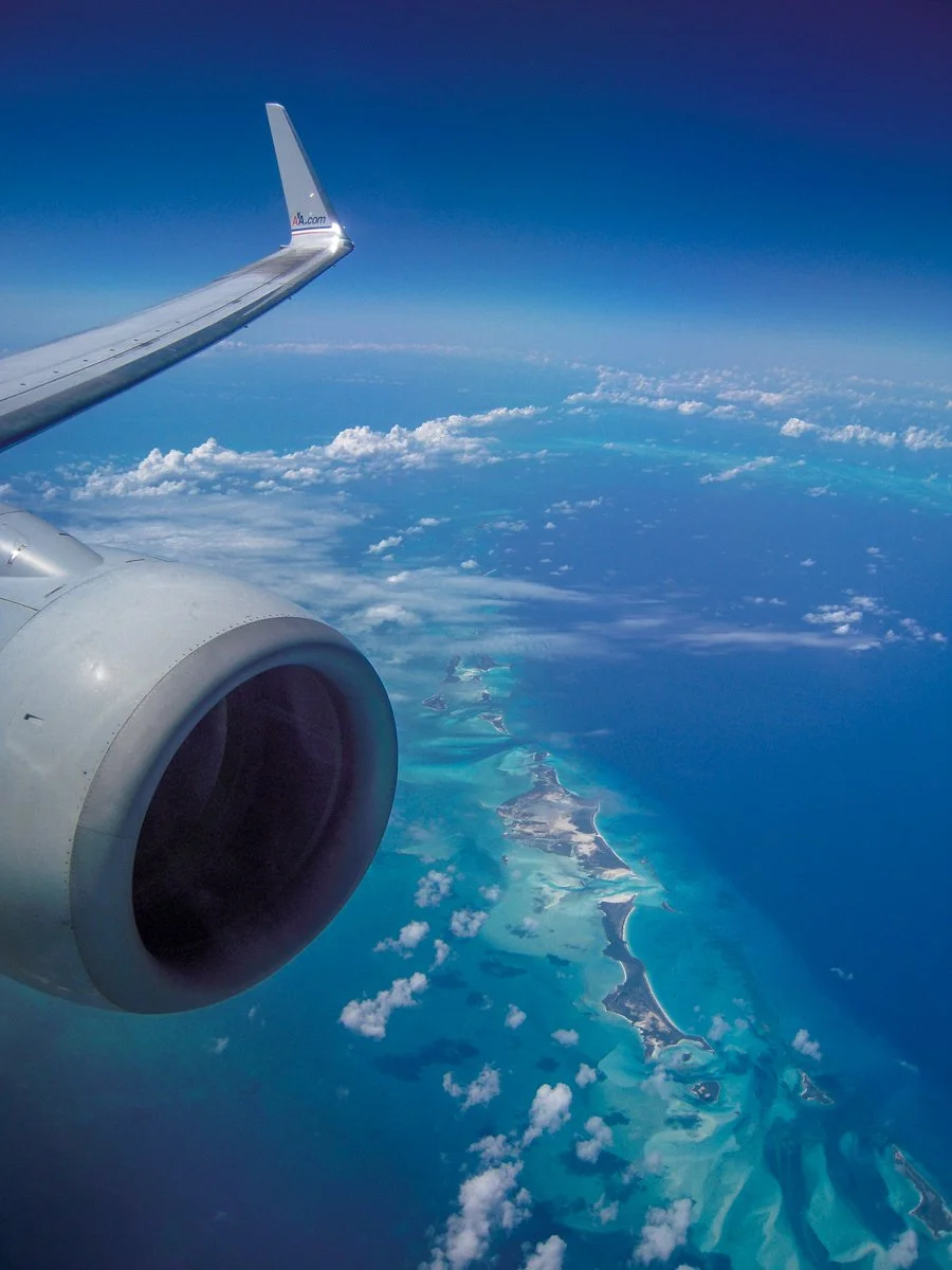 View from an airplane window showing the airplane wing and engine, with clouds and a coastline with islands below over the ocean.