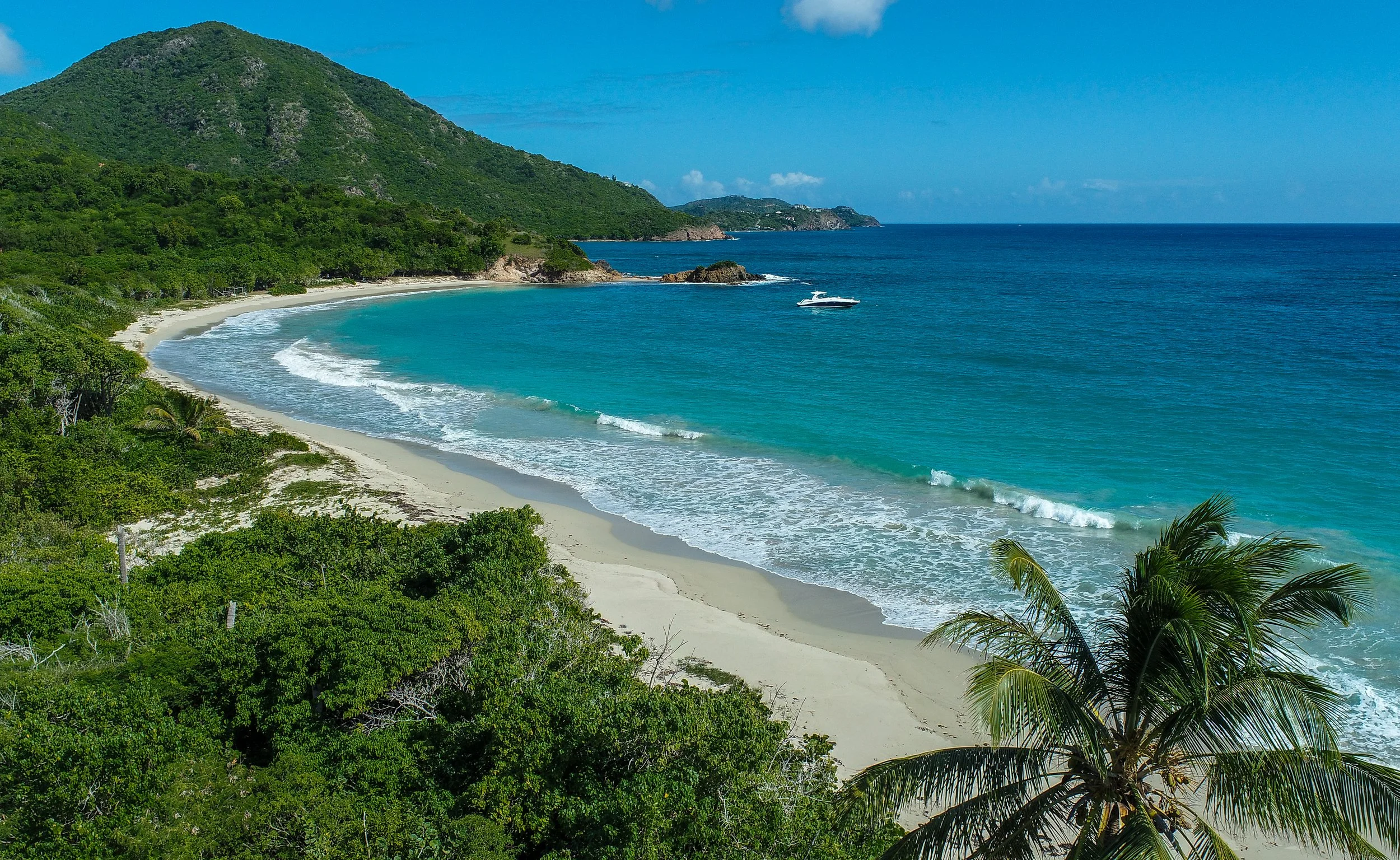 Scenic view of a tropical beach with white sand, turquoise water, lush green trees, and a mountain in the background.