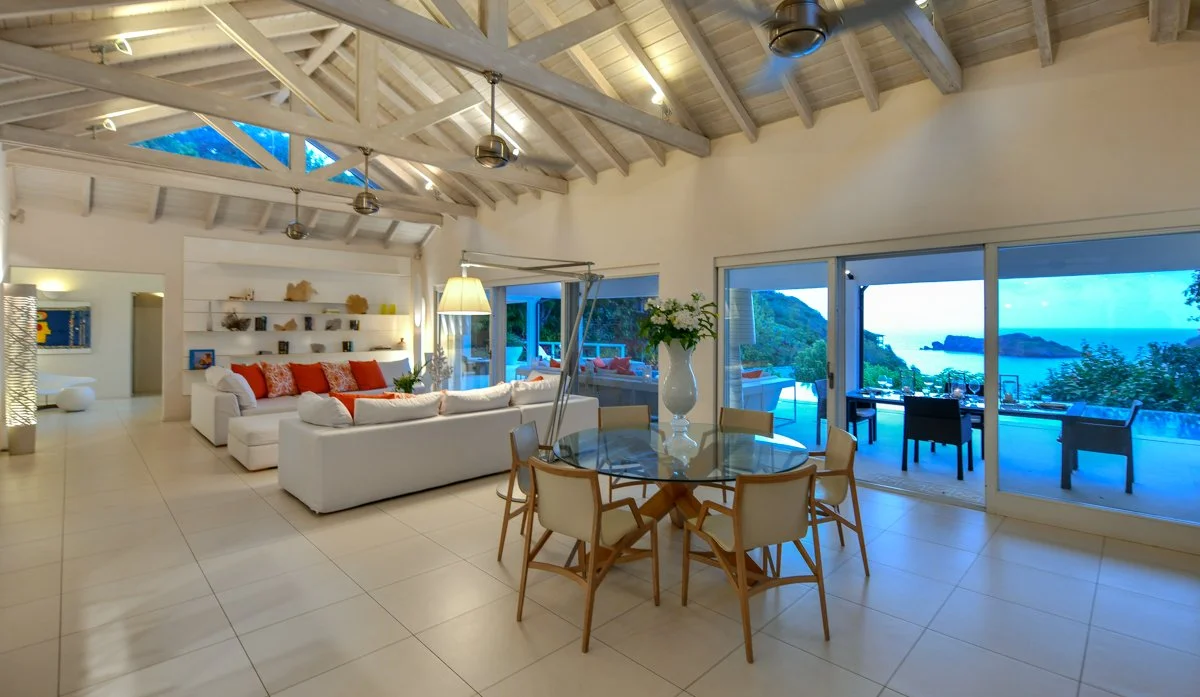 Living room with white sofas, a glass dining table with chairs, large windows, and a view of the ocean and outdoor patio.