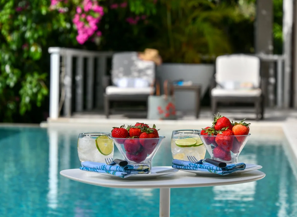 Two bowls of strawberries with lime slices and two glasses of water with lime slices on a white table near a swimming pool, with outdoor furniture and pink flowers in the background.