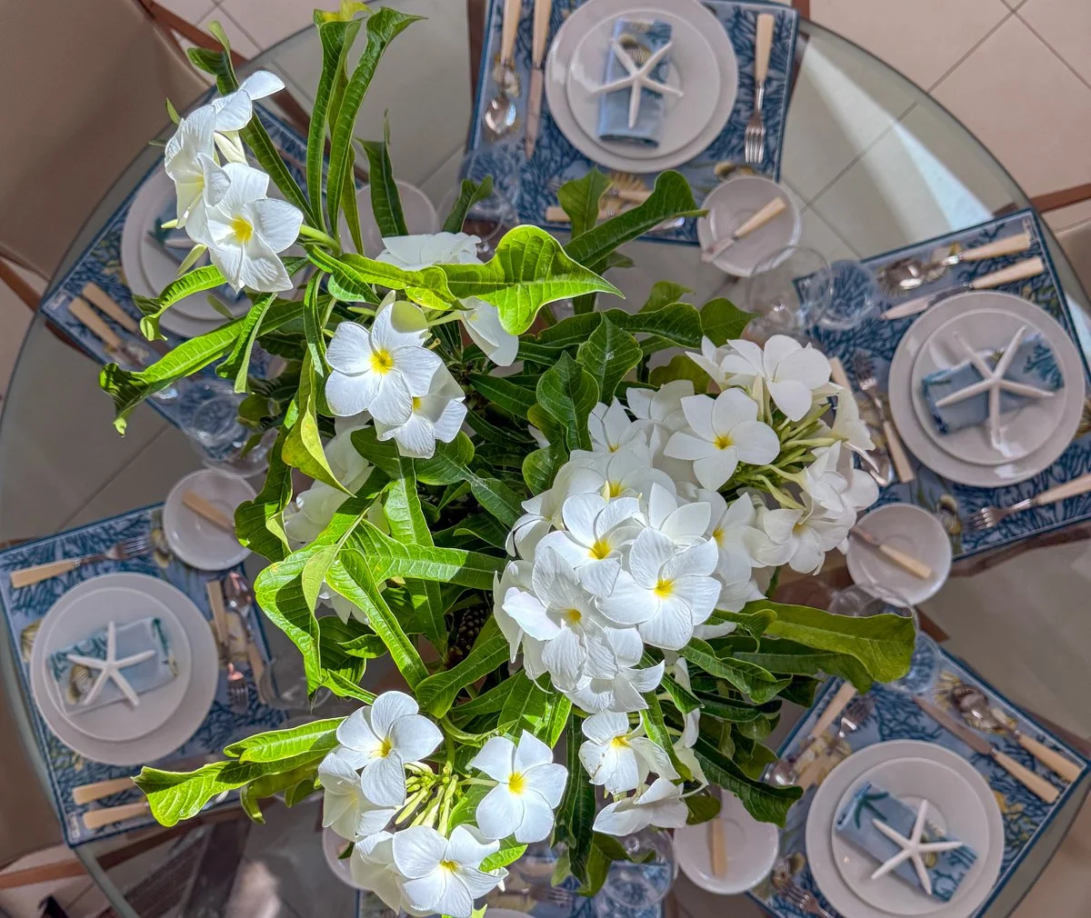 A dining table decorated with a large arrangement of white flowers and green leaves, set with white plates, starfish and blue napkins, and surrounded by chairs.