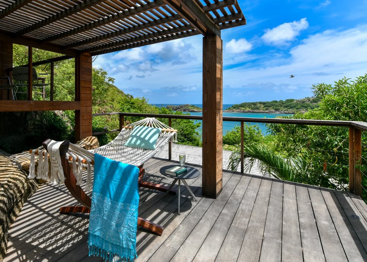A deck with a hammock, towels, cushion, and a side table with a drink, overlooking a scenic view of trees, ocean, and sky.