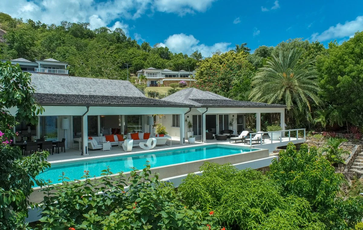 Modern house with covered patio, outdoor furniture, and a swimming pool surrounded by lush green trees and plants, set against a hilly landscape with a blue sky and scattered clouds.