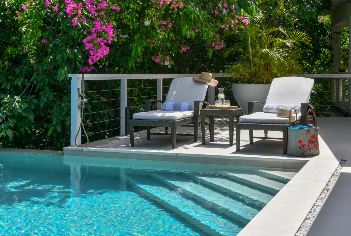 A backyard poolside patio with two white cushioned lounge chairs, one with a straw hat and striped towel, and a side table holding a pitcher and glasses. There's a large potted plant and blooming pink bougainvillea in the background.