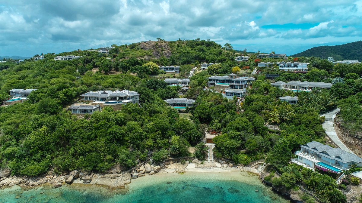 Luxury houses built on lush green hillside overlooking the ocean with a small beach at the bottom and a cloudy sky overhead.