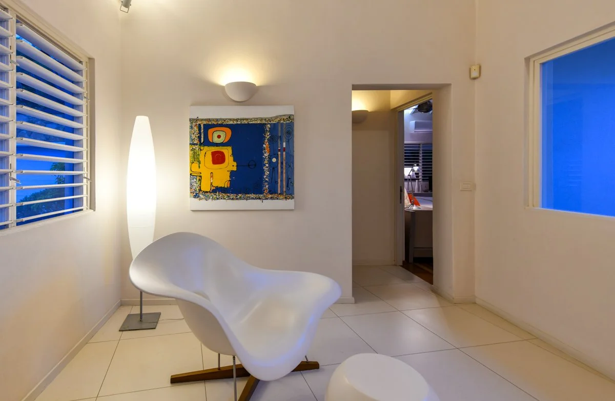 Modern living room with white walls, a white abstract chair, an oval white ottoman, and a vibrant abstract painting on the wall. There is a tall white floor lamp next to the chair. The room has two windows with white blinds, showing blue sky outside. Soft lighting creates a cozy atmosphere.