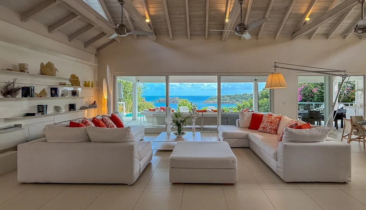 Living room with white sectional sofa, large windows showing ocean view, outdoor seating area on patio, and decorative elements like plants and lamps.
