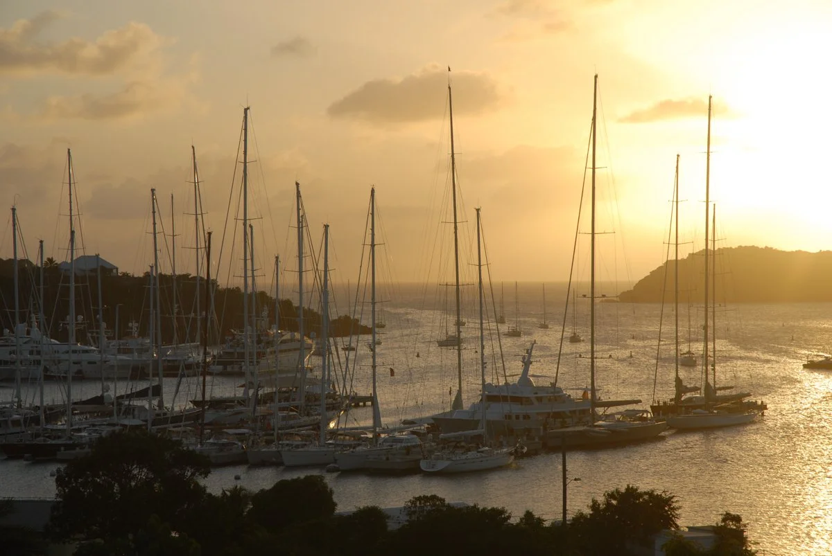 Yachts docked at a marina during sunset with hills and cloudy sky in background.
