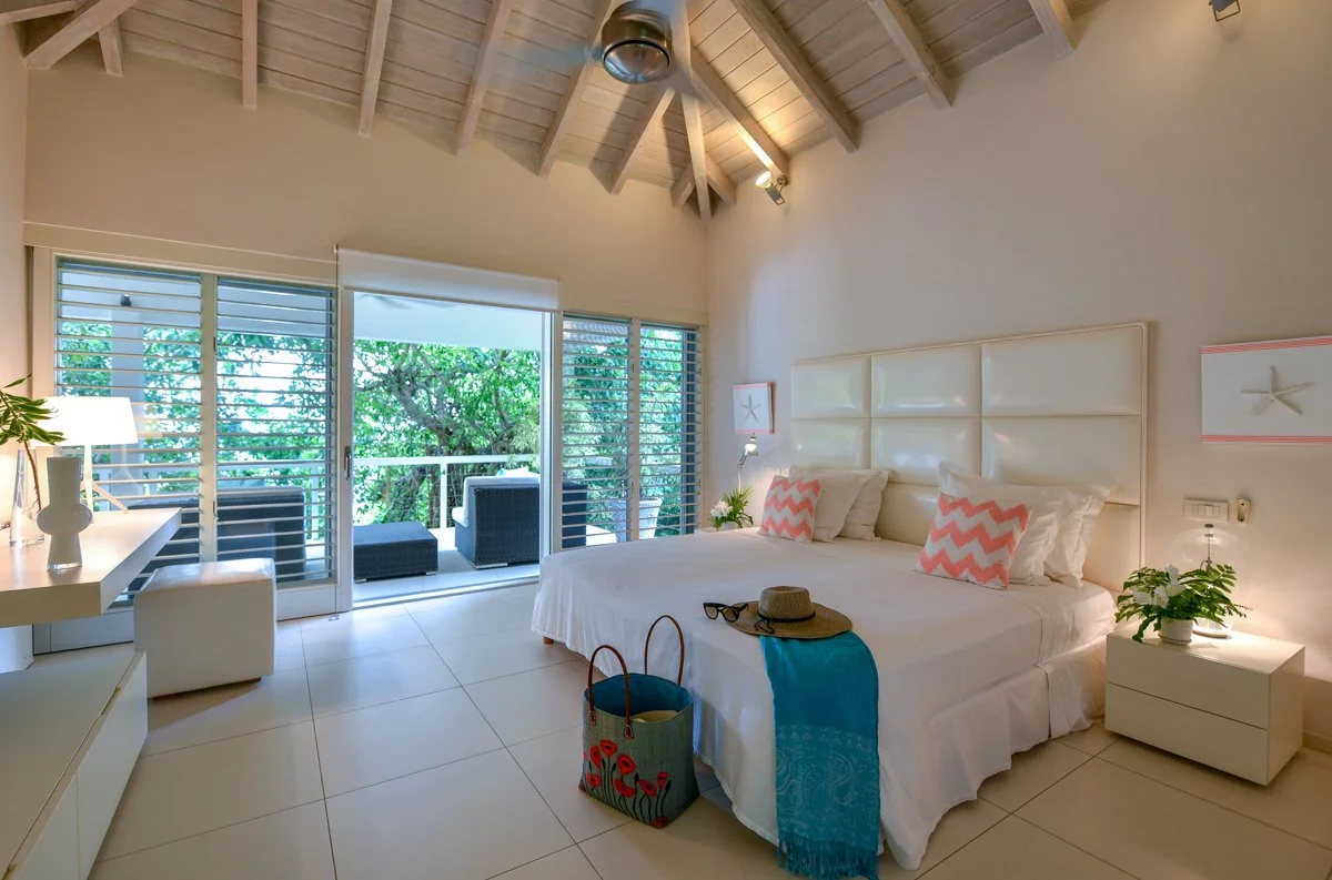 Bedroom with a large bed, pillows, a nightstand with a plant, a balcony with outdoor seating, and a high wooden ceiling.