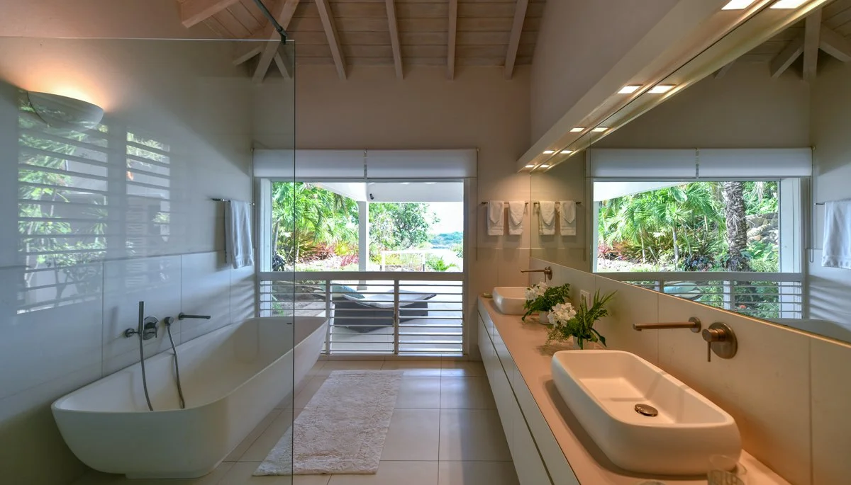 Bathroom with a large mirror, a bathtub next to a glass wall, two sinks, a vanity with plants and flowers, towels on the wall, and a view of lush greenery and a boat outside.