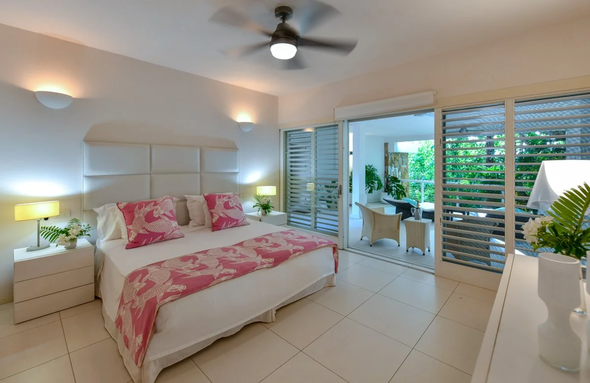 A bedroom with a white bed, pink and white pillows, and a pink blanket with white bird patterns, two nightstands with lamps and plants, a ceiling fan, and sliding glass doors leading to a balcony with outdoor seating and greenery.