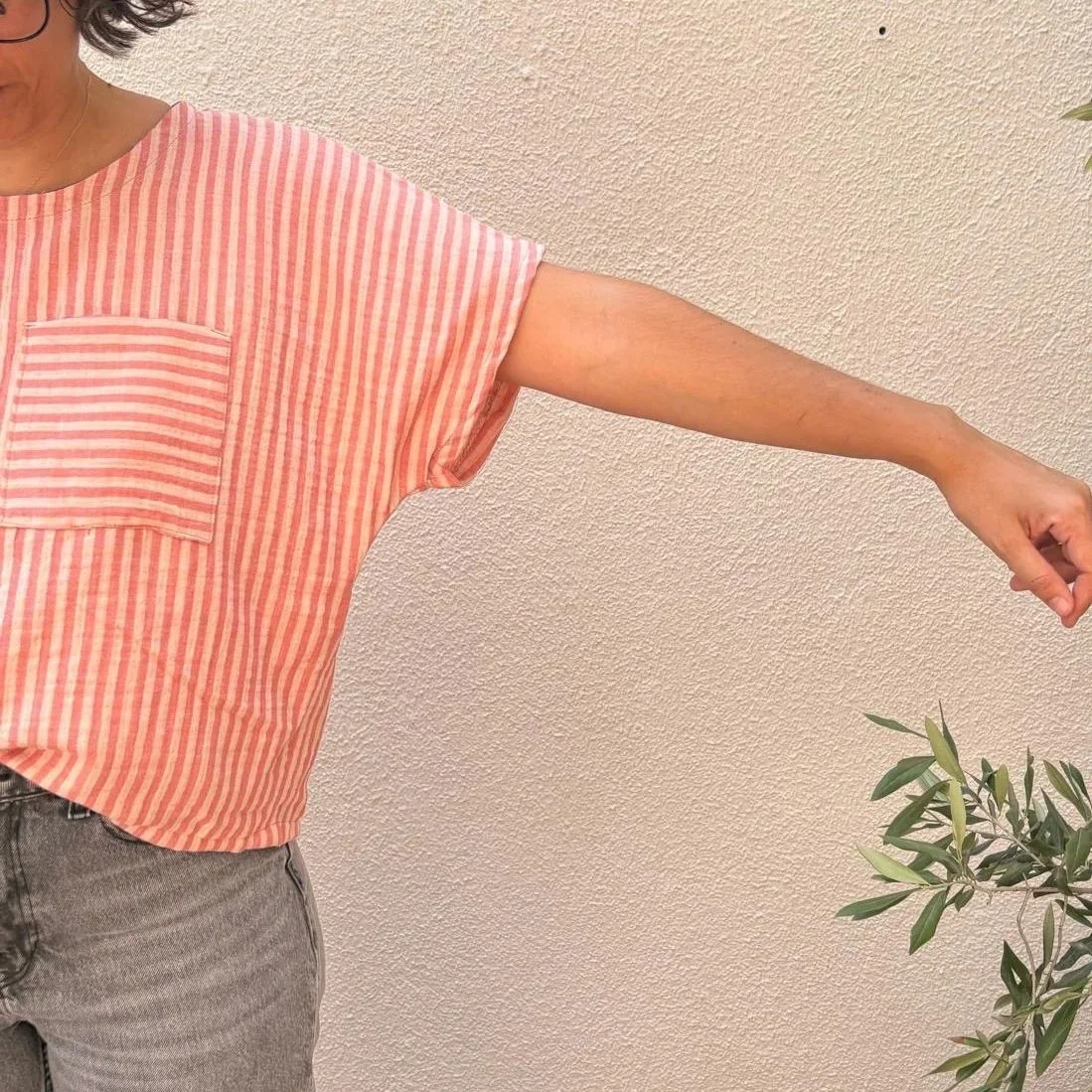 Persona con camiseta de rayas rojas y blancas, dando un abrazo y estirando su brazo hacia la derecha, fondo de pared beige y planta en la esquina inferior derecha.