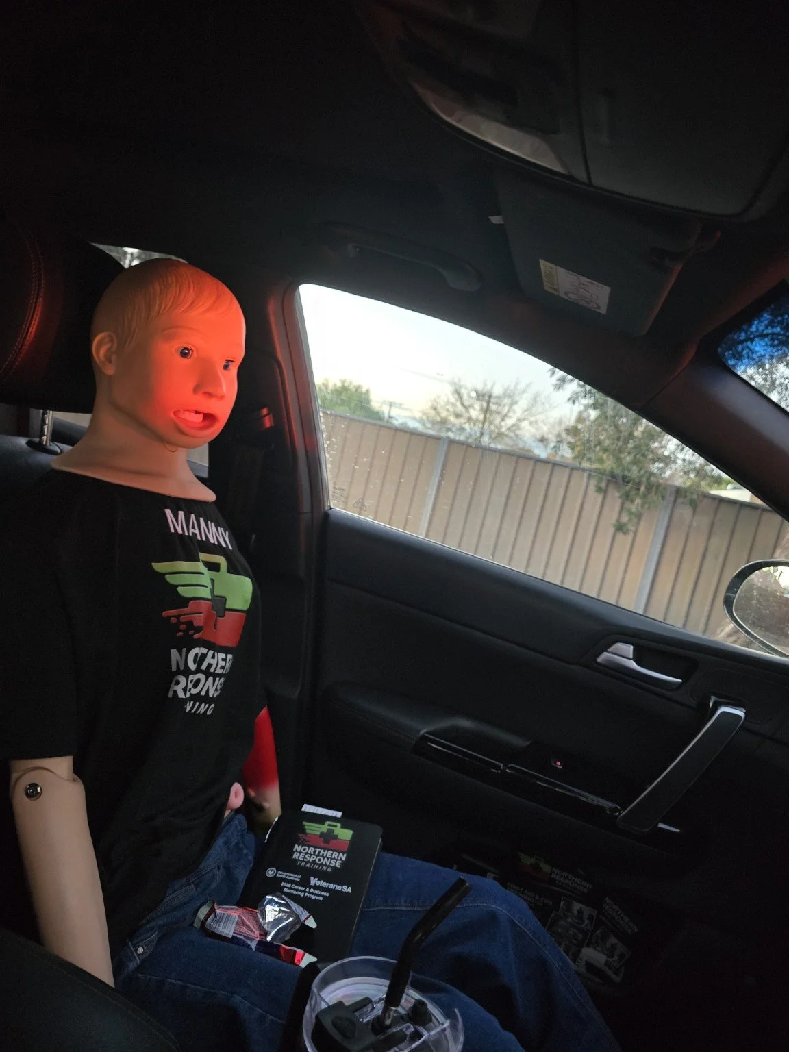 A mannequin with a realistic child's face is seated in the driver's seat of a car. The face appears surprised or shocked, and the mannequin is wearing a black T-shirt with a racing logo. The car's interior and a fence Manny ready for the road