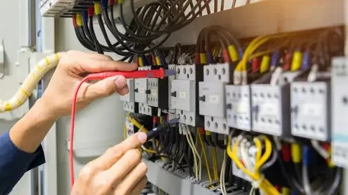 A person using a multimeter to test electrical wiring inside an electrical panel.