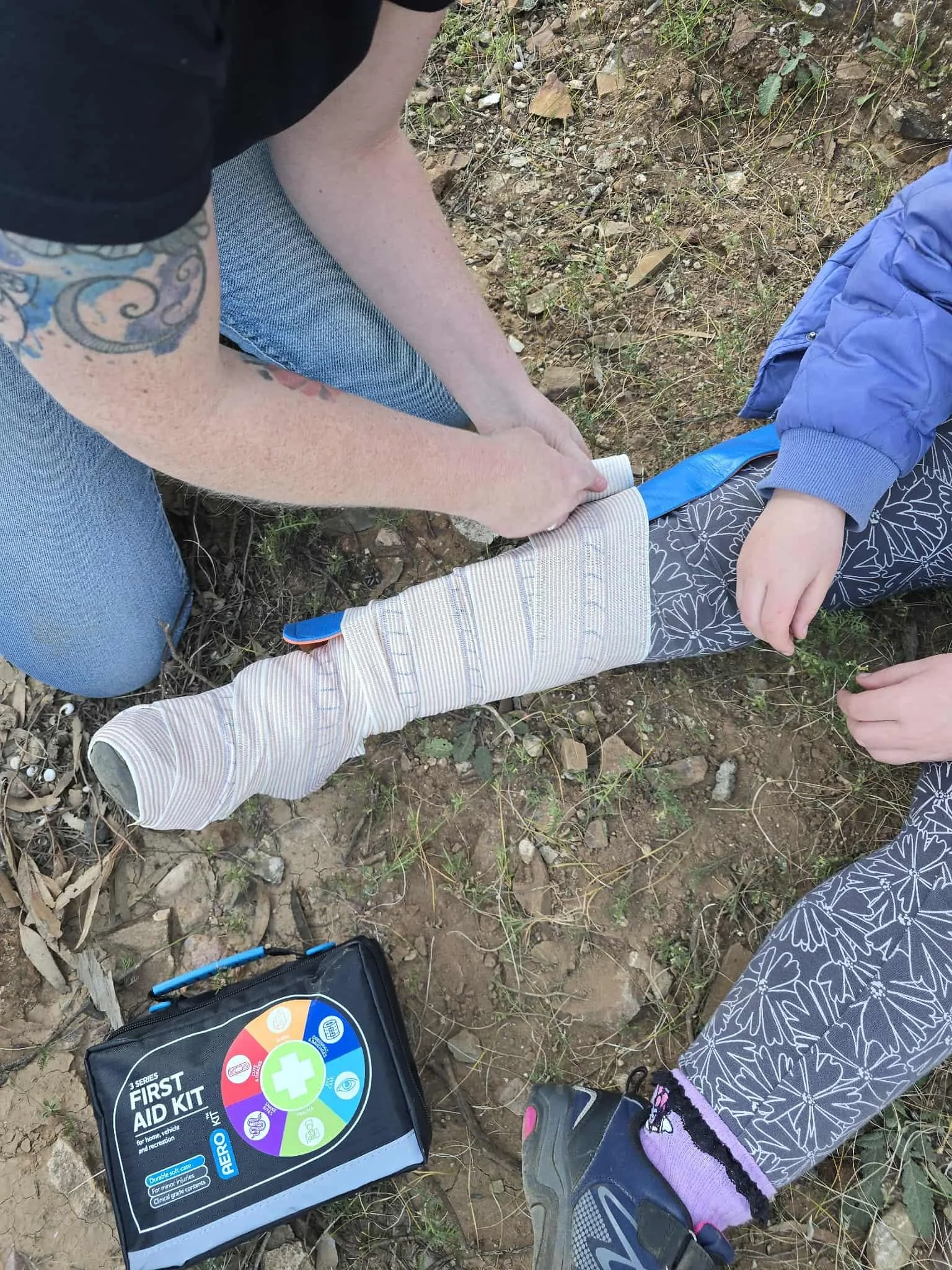 A person in a black shirt kneels on dirt ground, applying a tourniquet to a child's leg, with a first aid kit nearby.