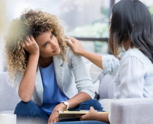 Two women having a supportive conversation in a bright, modern setting with a window in the background. One woman with curly hair appears distressed, while the other woman is listening and offering comfort.