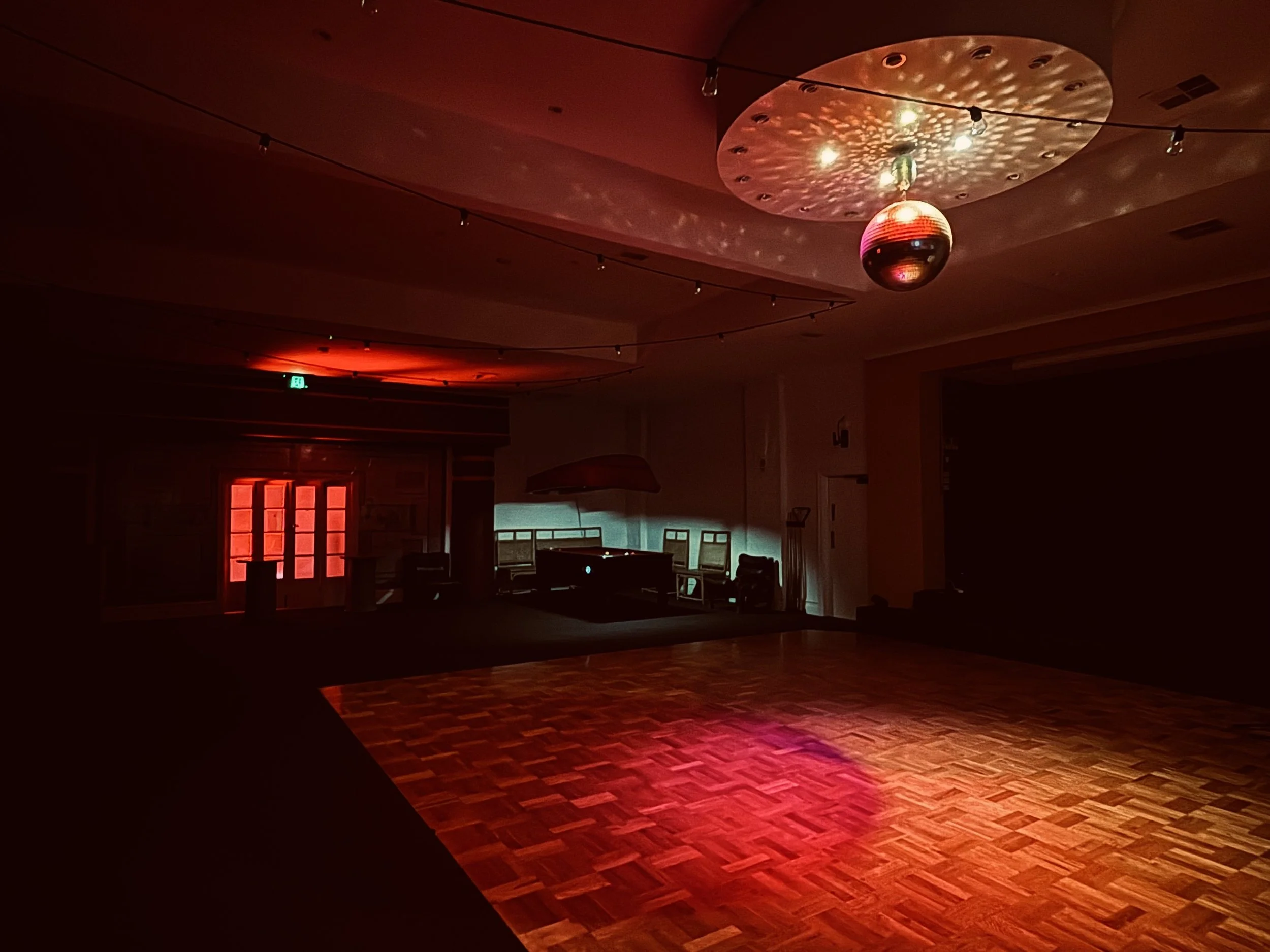 Empty dimly lit dance floor with a disco ball hanging from the ceiling, colorful lights casting reflections, and a DJ booth with speakers in the background.