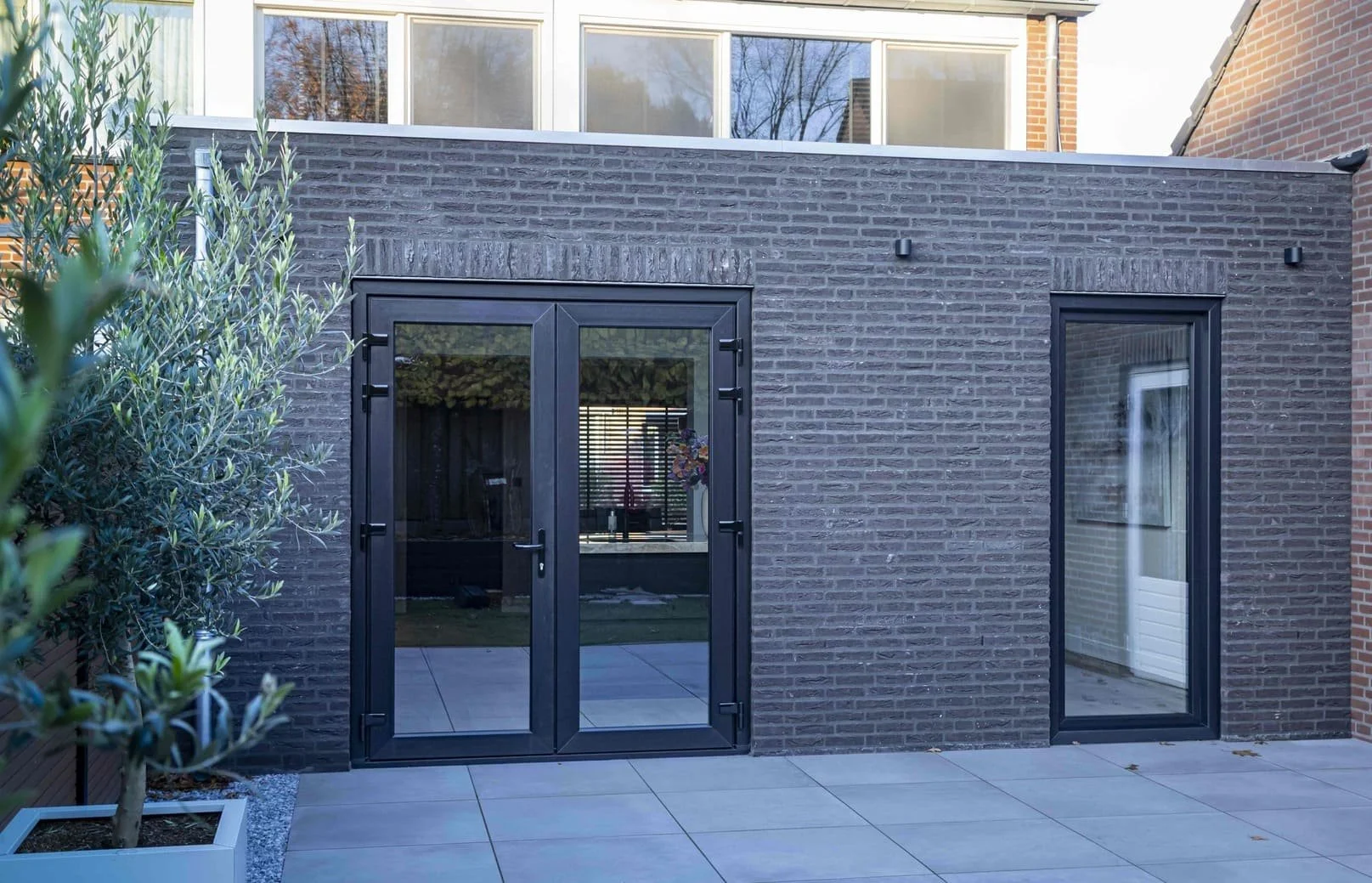 Moderne gevel met zwarte metalen deuren en ramen, een terras met grote tegels, en een boom in een witte bloempot naast de gevel.