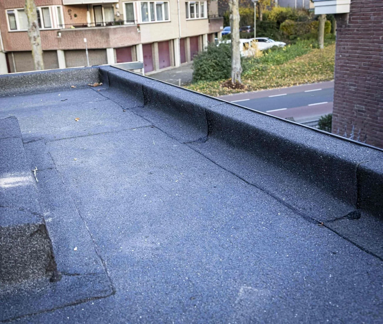 Close-up van een zwart geschilderd plat dak met kiezels en een opstaande rand, met een woonwijk op de achtergrond, inclusief bomen, stoep en straat.