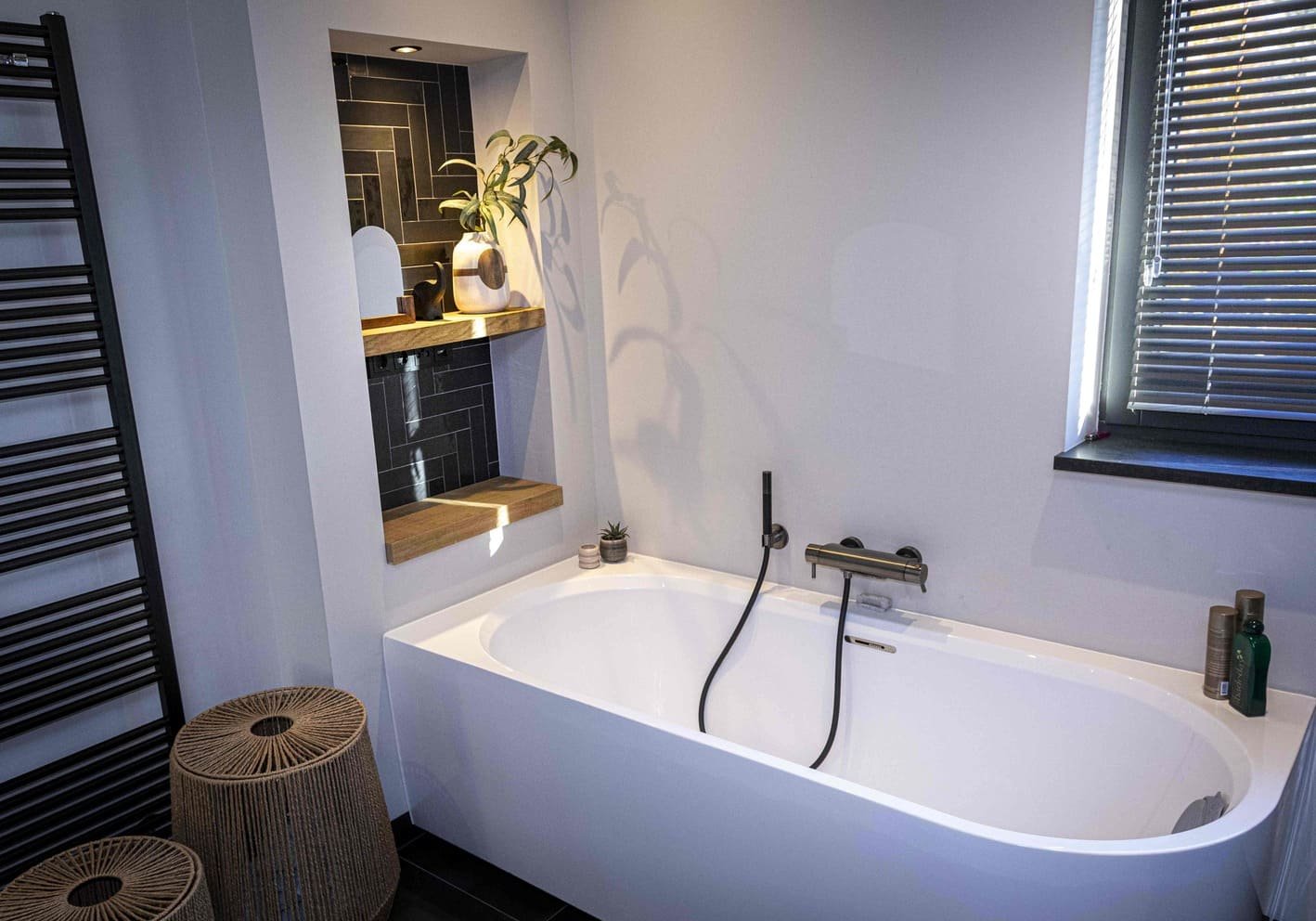 Moderne badkamer met wit vrijstaand bad, zwarte muurtegels in nis met decoratieve vazen en planten, en een raam met jaloezieën.