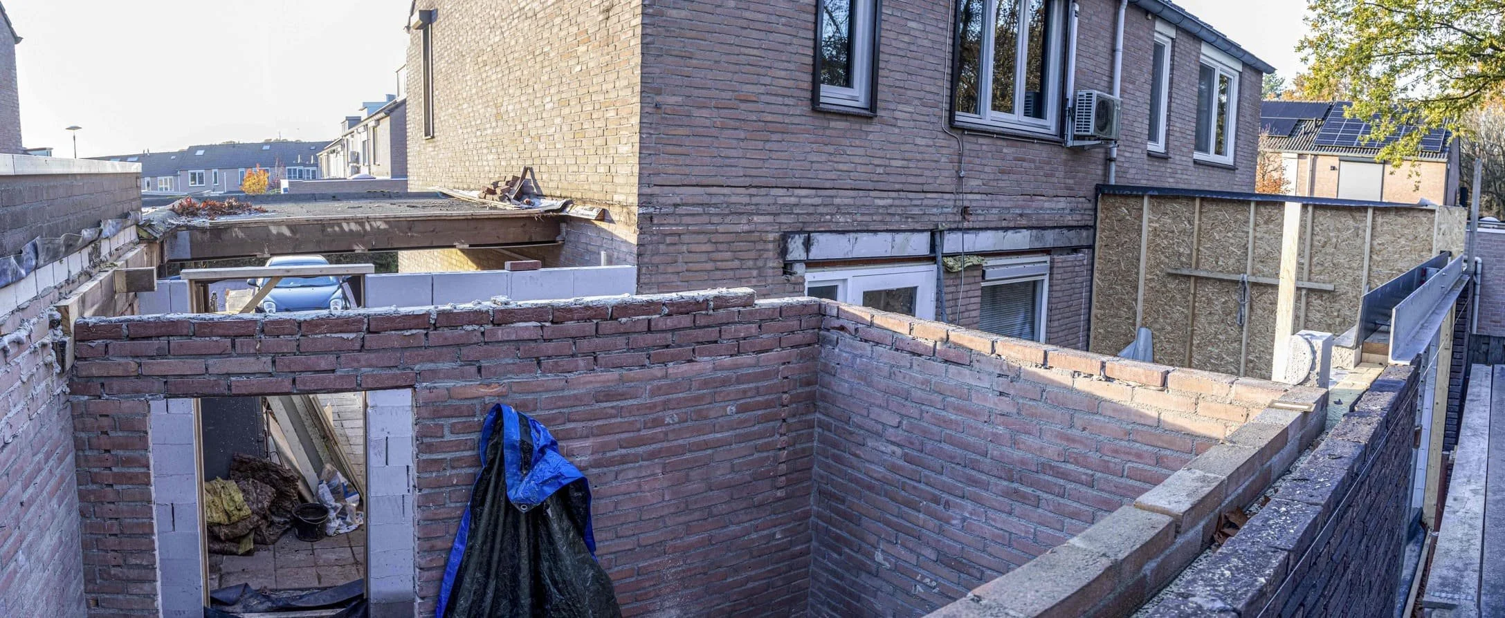 Bouwplaats met gedeeltelijk opgebouwde bakstenen muren, houten steunbalken en bouwmaterialen, in een woonwijk met bestaande huizen.