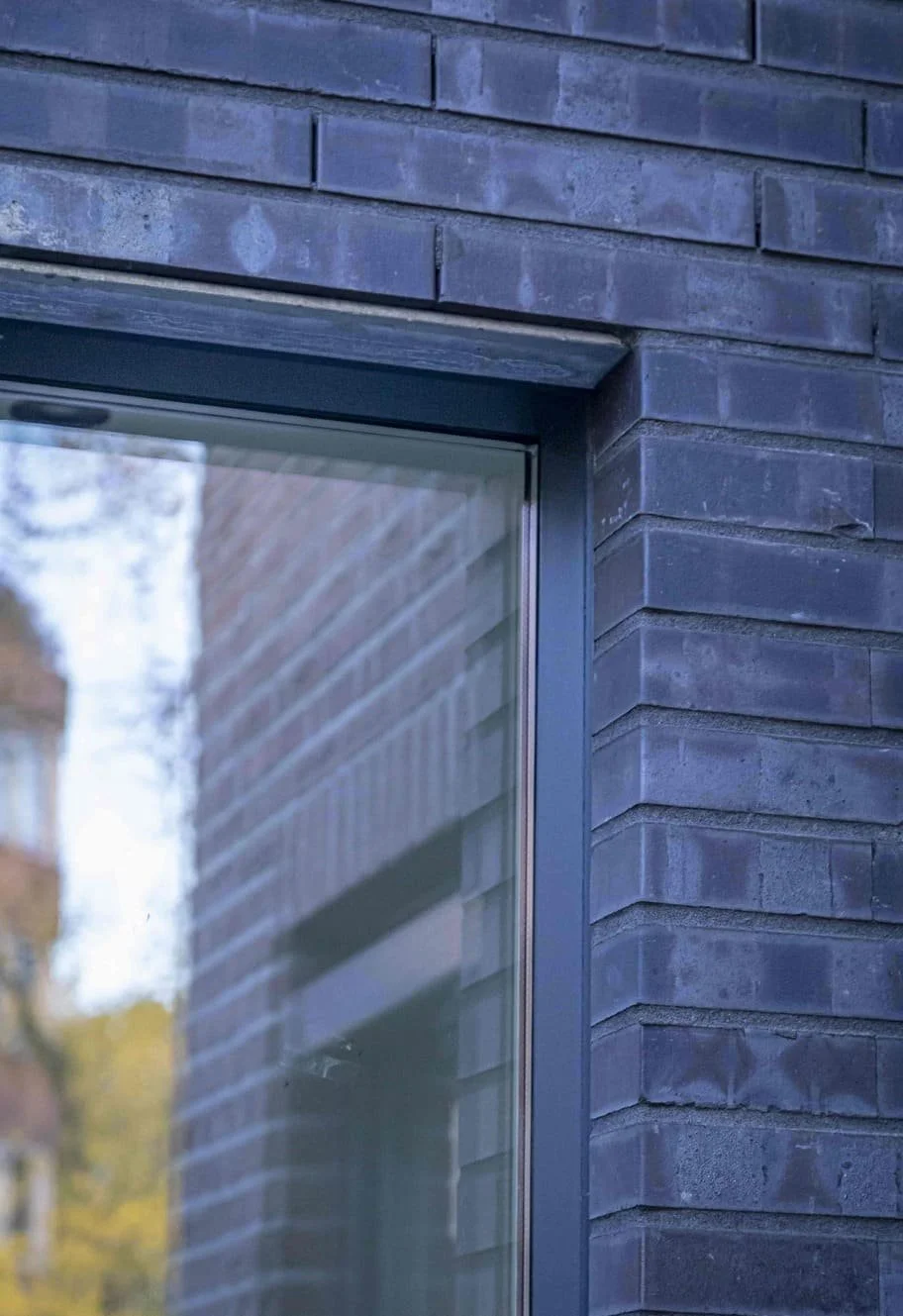 Close-up van een raam in een muur van donkergrijze bakstenen met een reflectie van een boom en de lucht.
