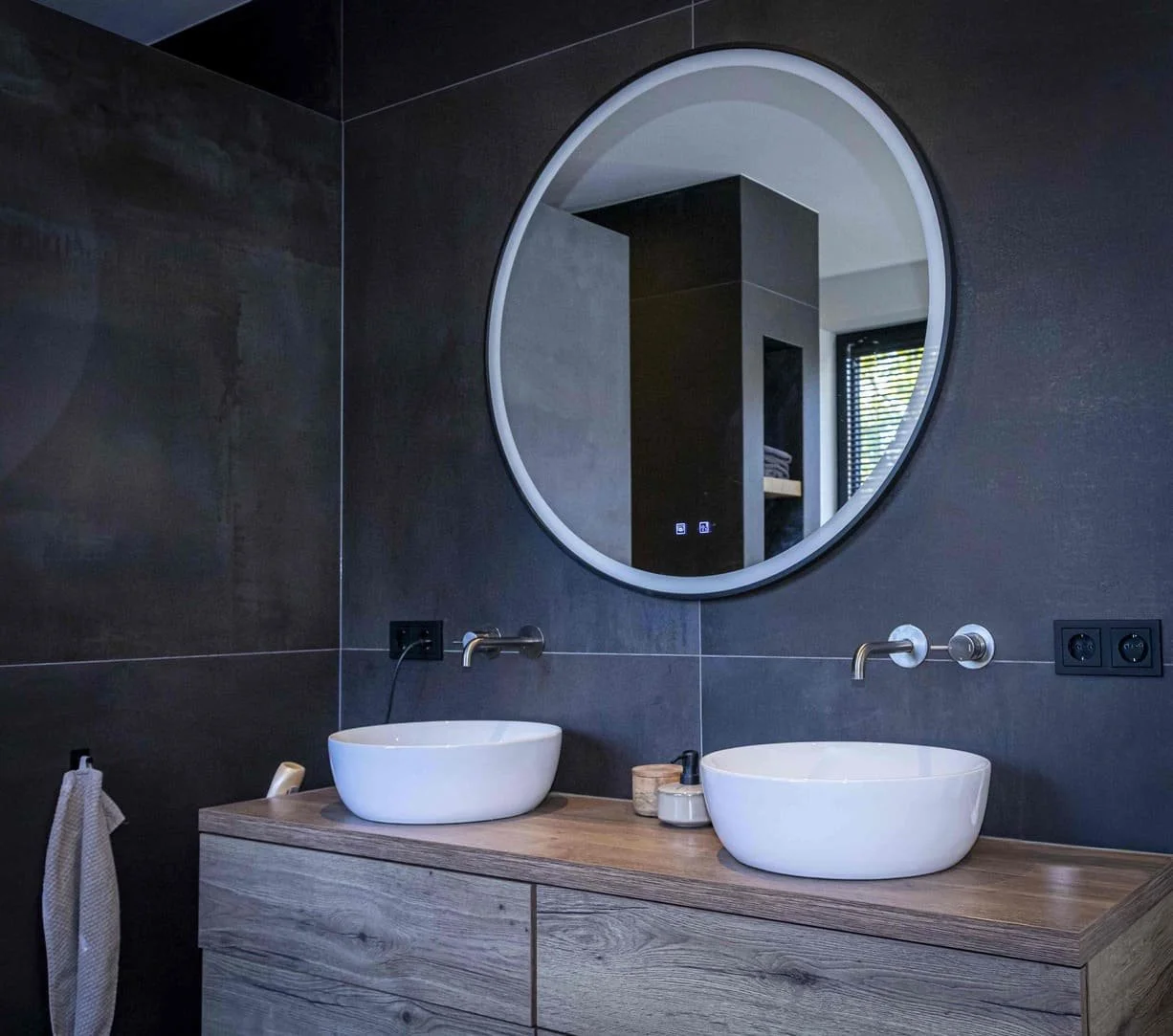 Moderne badkamer met donkergrijze tegels, ronde spiegel, dubbele waskommen op houten wastafel, wandfittingen en stopcontacten.