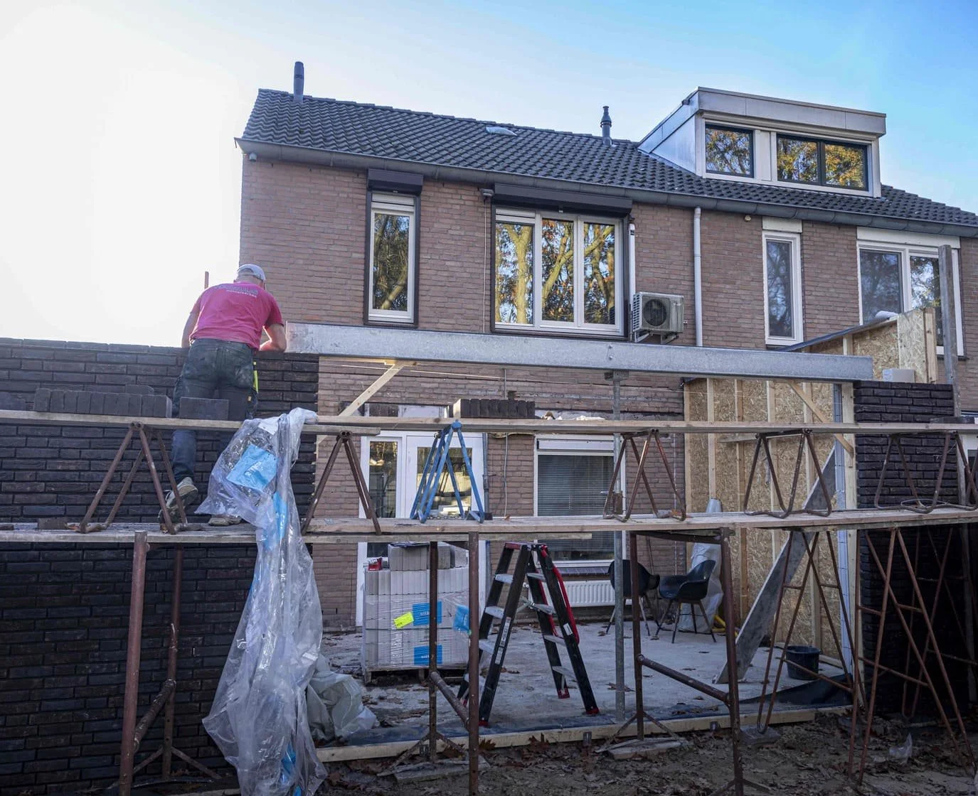 Een bouwvakker werkt aan de buitenkant van een huis, hij staat op een steiger en metselstenen worden gelegd op de muur.