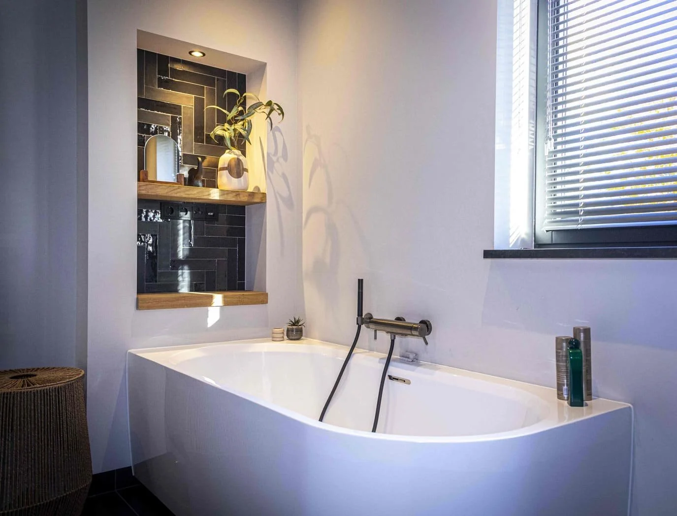 Moderne wit badkuip met zwarte tegels en houten planken, decoratieve planten en badproducten voor een ontspannende badkamer.