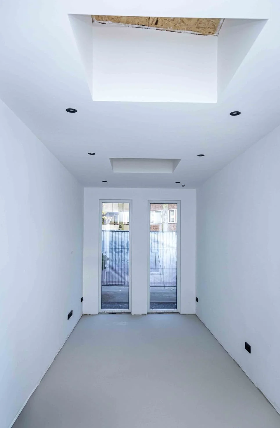 Een binnentrap zonder vloer, plafond met uitsparingen, en twee grote ramen achterin de kamer, klaar om afgewerkt te worden.