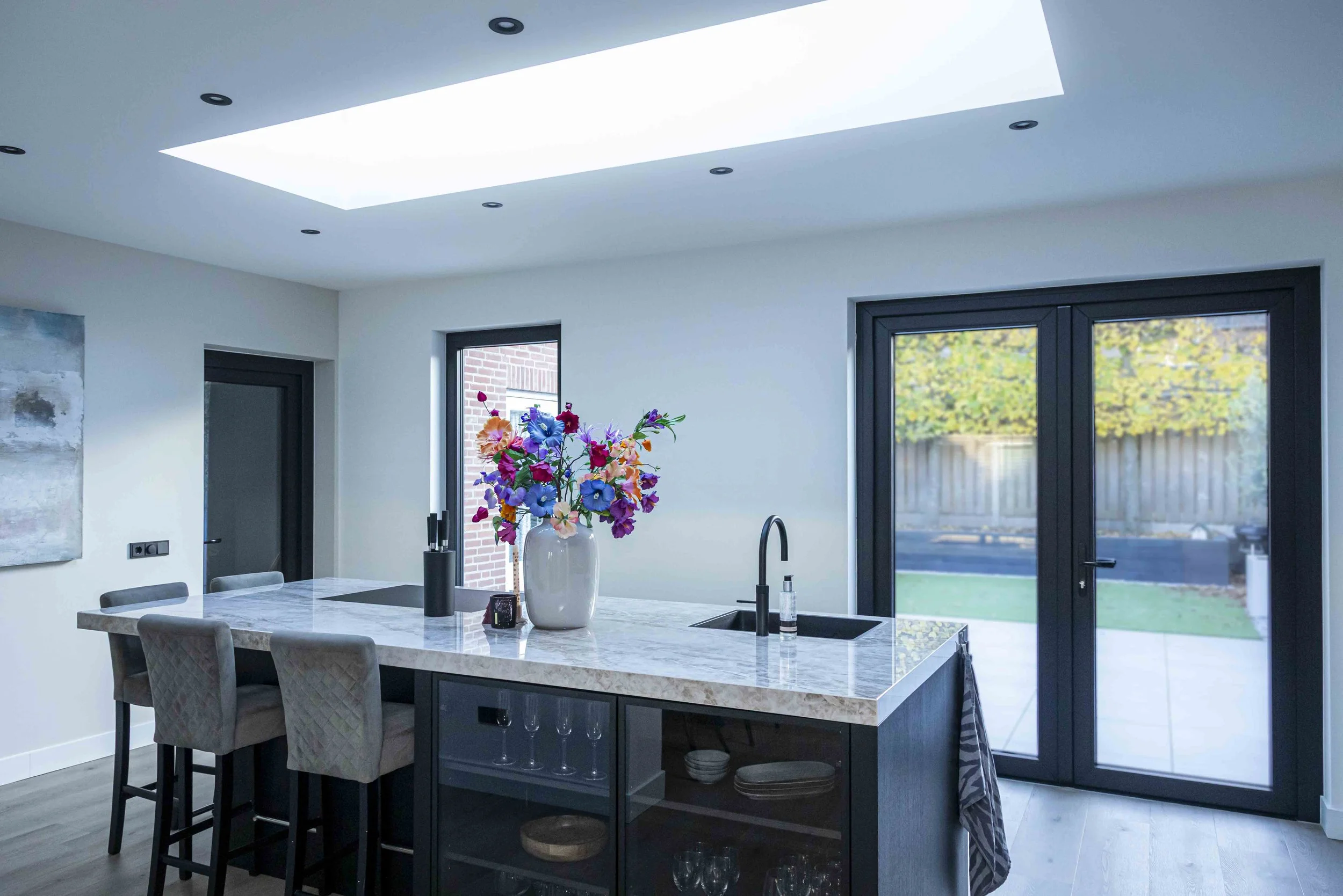 Een moderne keuken met een marmeren kookeiland, een grote vaas met kleurrijke bloemen, zwarte accenten en schuifpuien die uitkomen op een achtertuin met houten schutting en bomen.