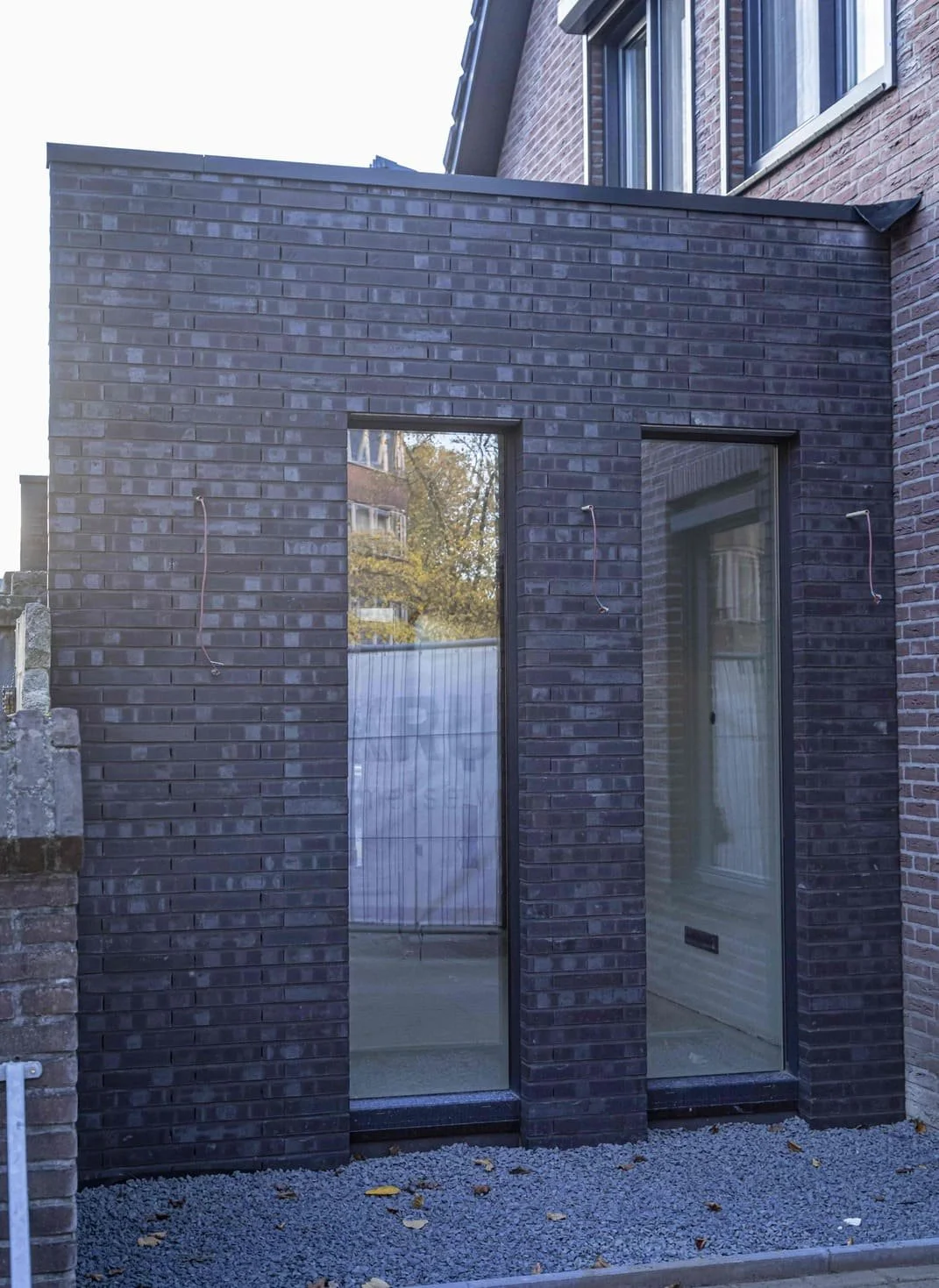 Nieuw gebouw met donkere bakstenen gevel en grote ramen, aangelegd terras met grind voor de ingang.