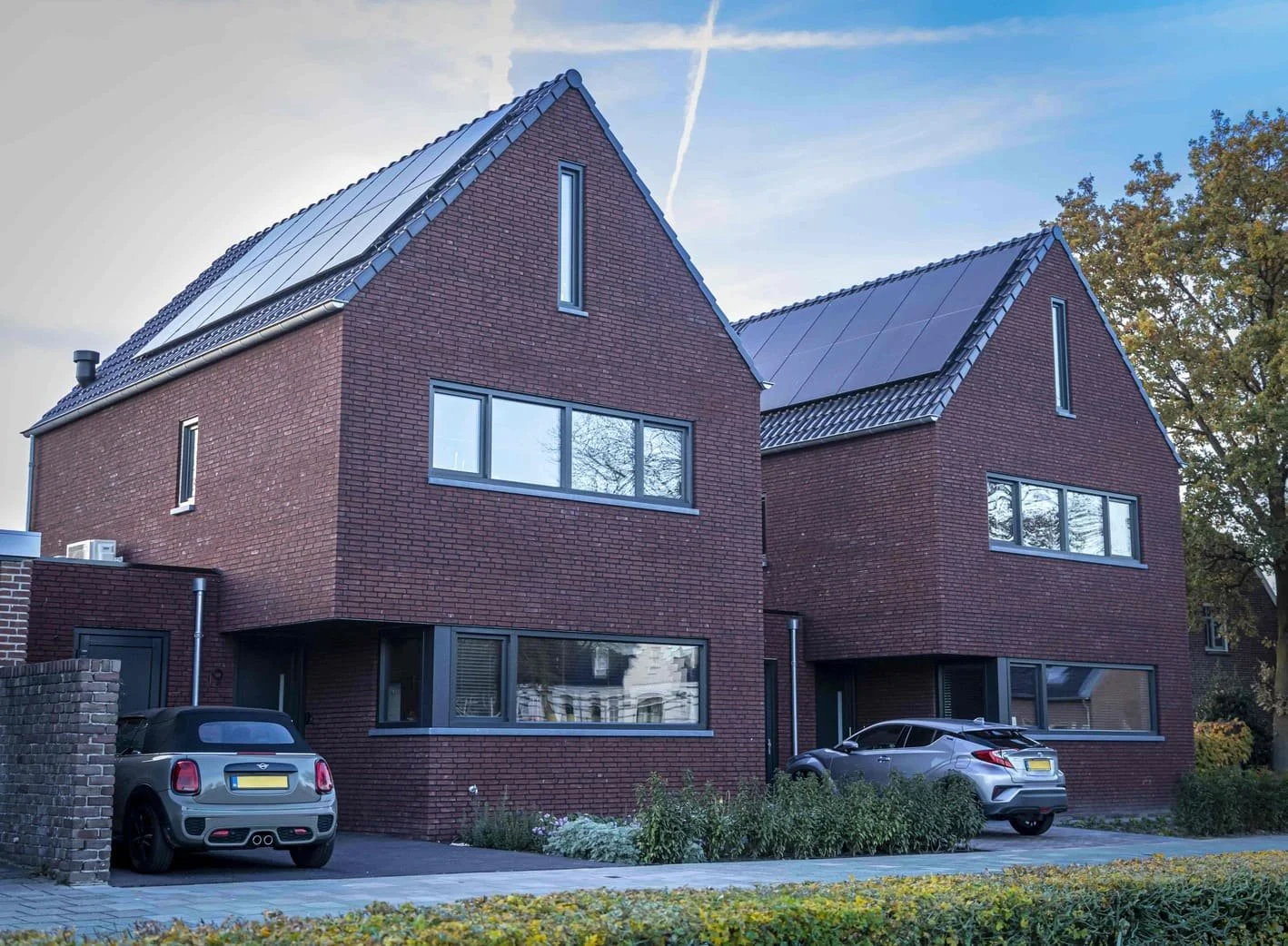 Twee moderne bricken huizen met grote ramen, parkeerruimte met twee auto's, en een tuin met planten, onder een blauwe lucht.